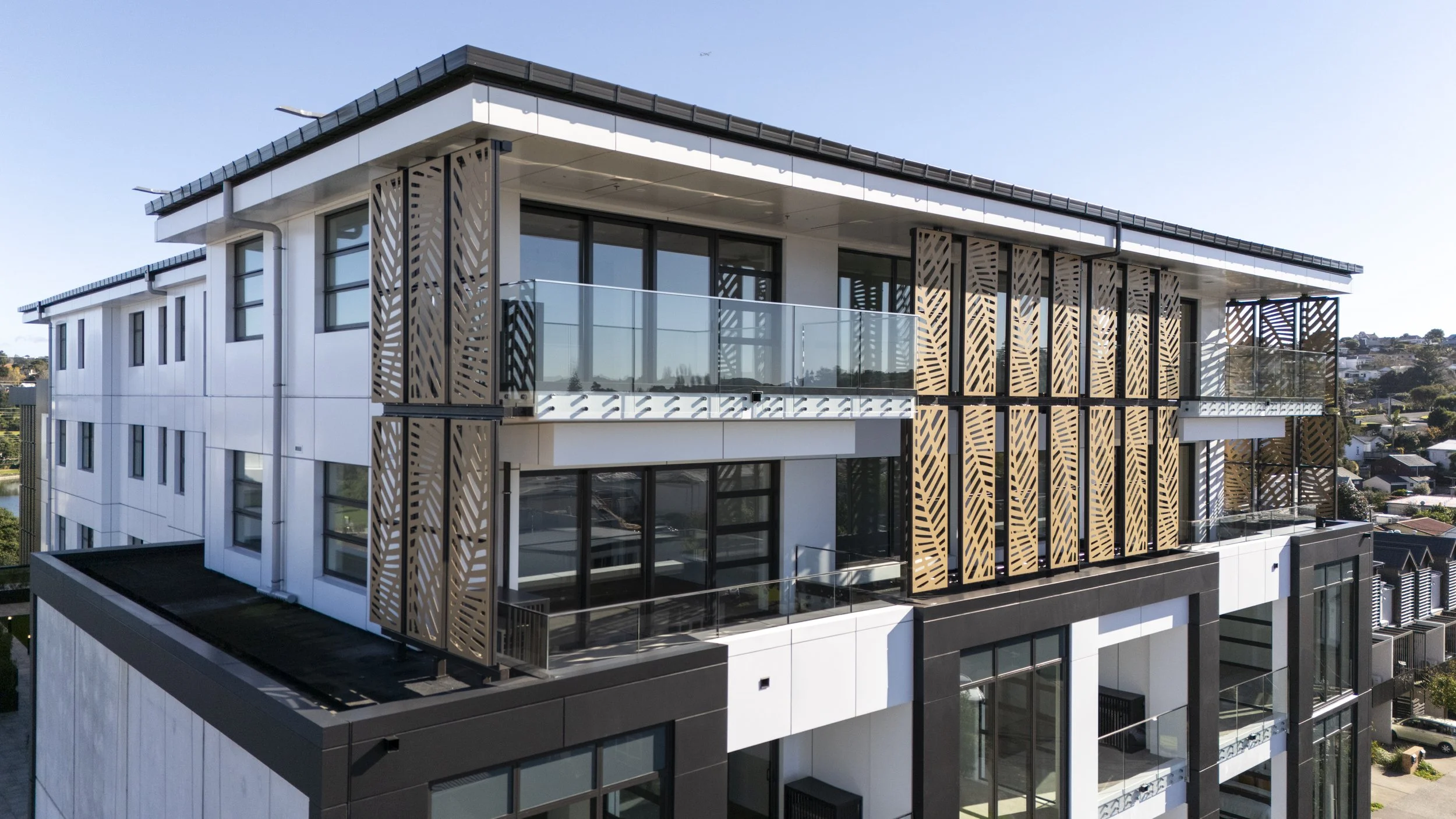 A modern multi-story apartment building with a white and black exterior, glass balcony railings, and decorative wooden panels, situated in a suburban neighborhood.