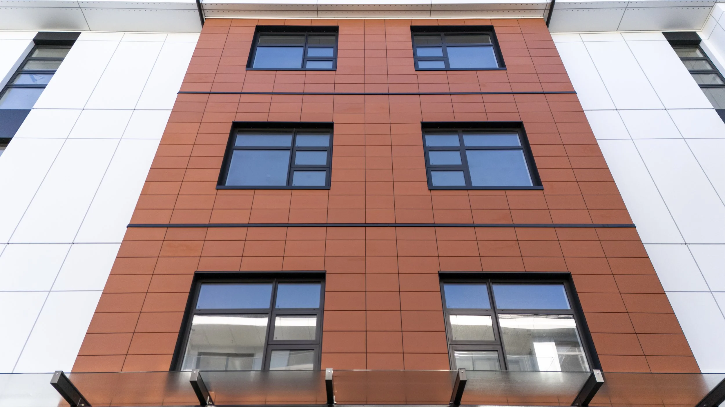 Close-up of a modern building's facade featuring a central section with orange tiles and black-framed windows, flanked by white surfaces with vertical window stripes.
