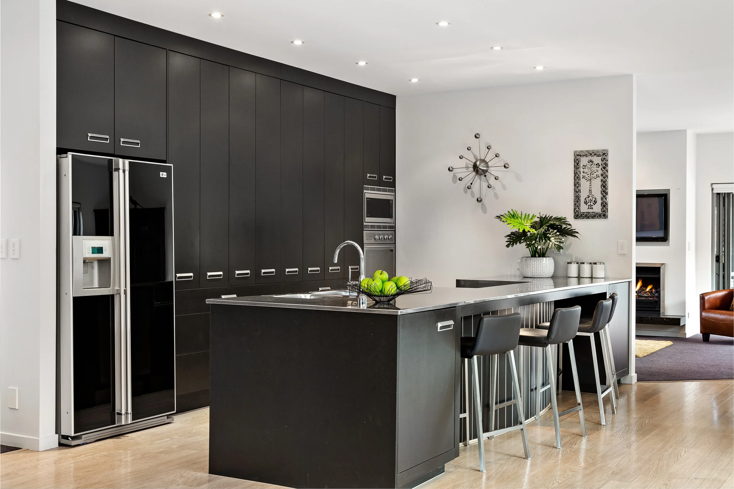Modern kitchen with black cabinets, stainless steel refrigerator, built-in microwave, kitchen island, barstools, decorative wall art, potted plant, and a living area with a fireplace and a small TV.