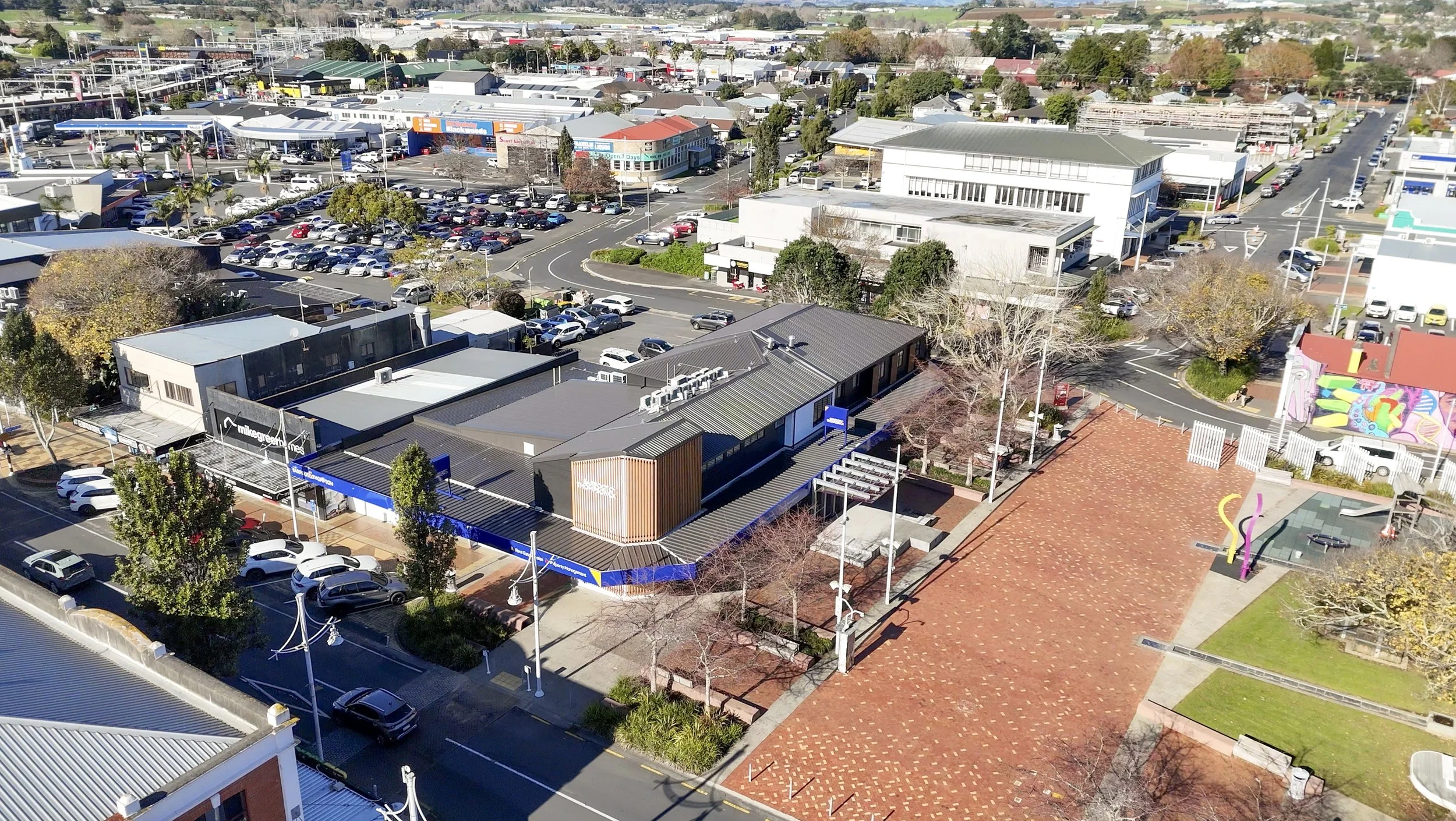 Aerial view of a commercial shopping area with parking lots, retail stores, some trees, and a large open brick-paved plaza area.