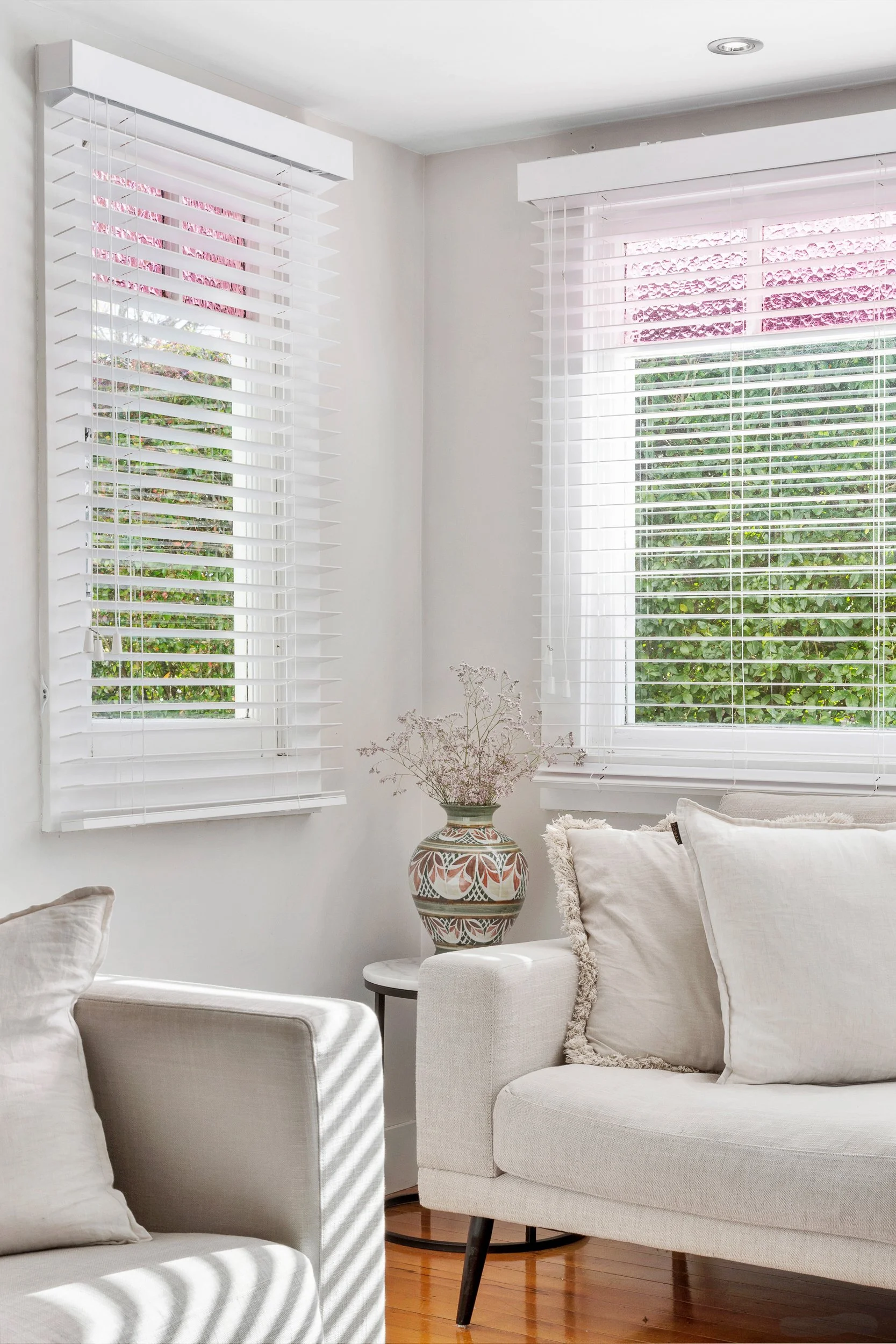 Living room with large white armchairs, white walls, and two windows with white blinds. A decorative vase with plant is placed on a white table between the chairs. Light creates shadow stripes on the furniture.
