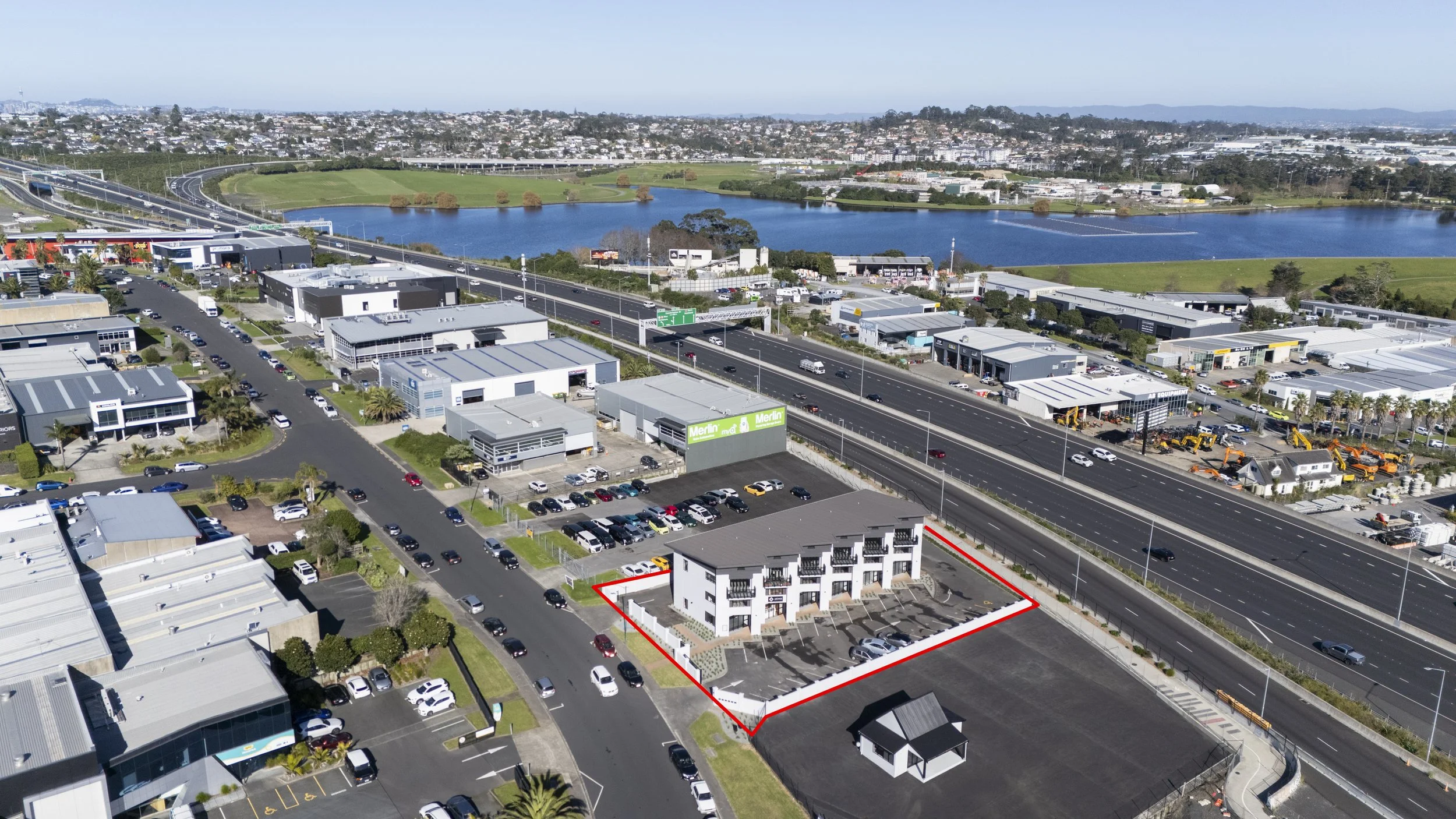 Aerial view of a commercial area with a parking lot, various buildings, a highway, and a lake in the background, with residential and industrial areas beyond.