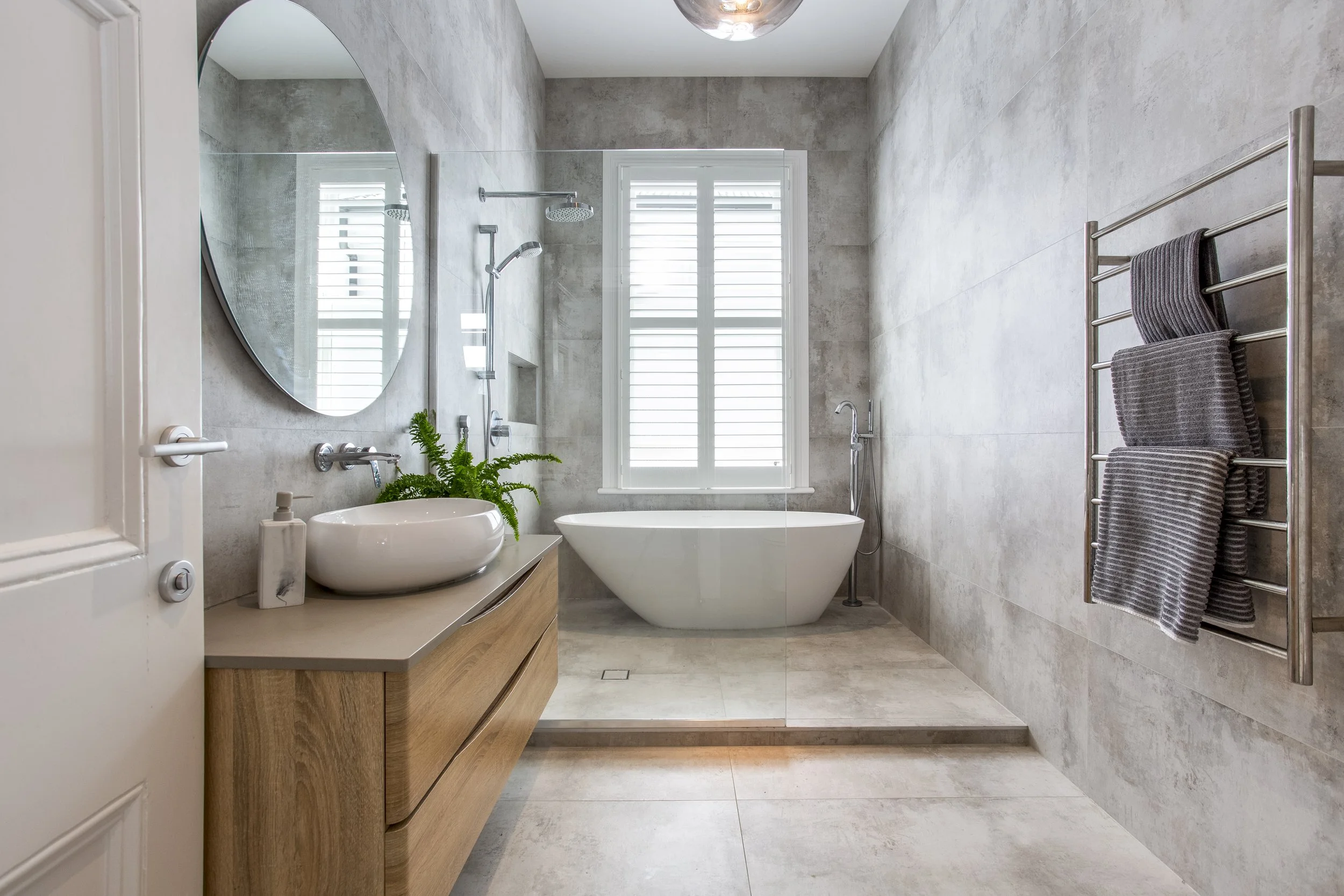 Modern bathroom with a freestanding bathtub, gray tiled walls, a large window with shutters, a mounted showerhead, a wooden vanity with a round vessel sink, a mirror, a potted plant, and a towel rack with dark gray towels.