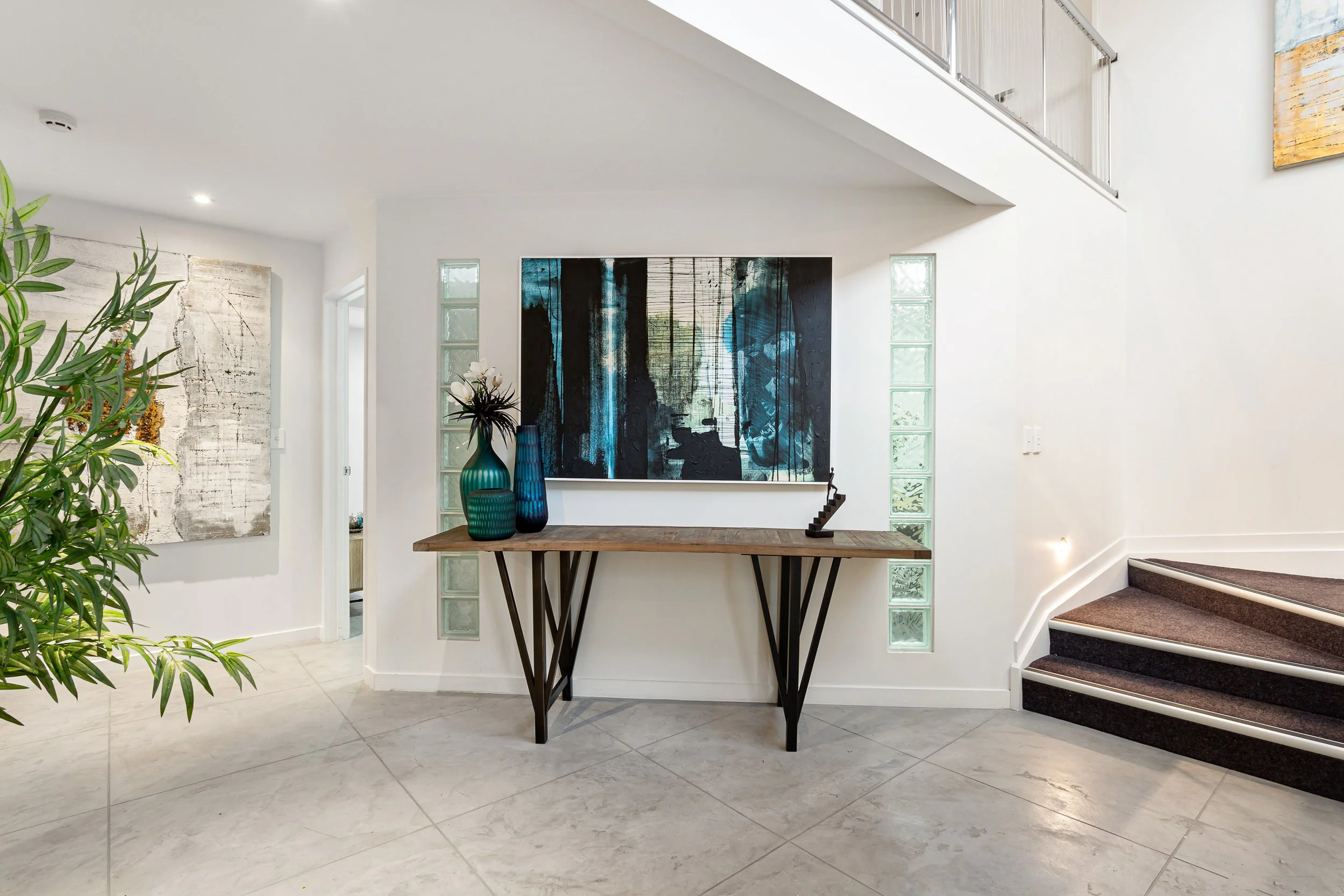 Modern interior with white walls, staircase with dark carpet, large sliding glass door, and abstract artwork on the wall. Decorative vases and plant on a wooden table in the entrance area.