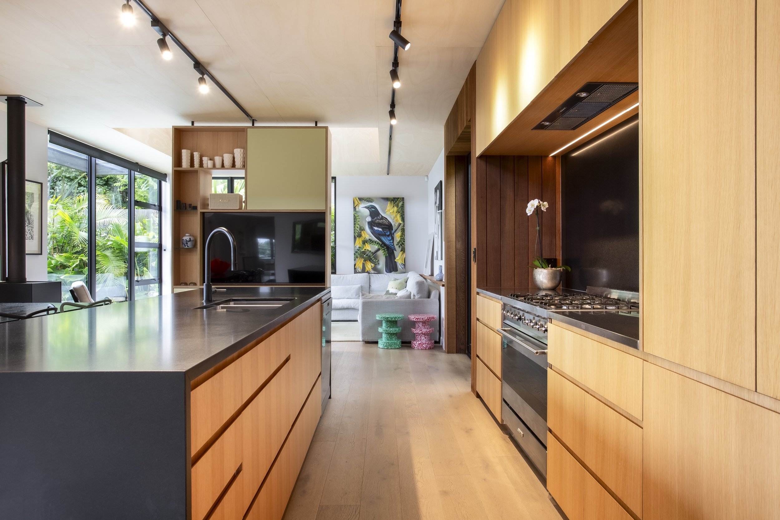 Open-concept kitchen with wooden cabinets, black countertops, stainless steel oven, and large windows overlooking a garden.