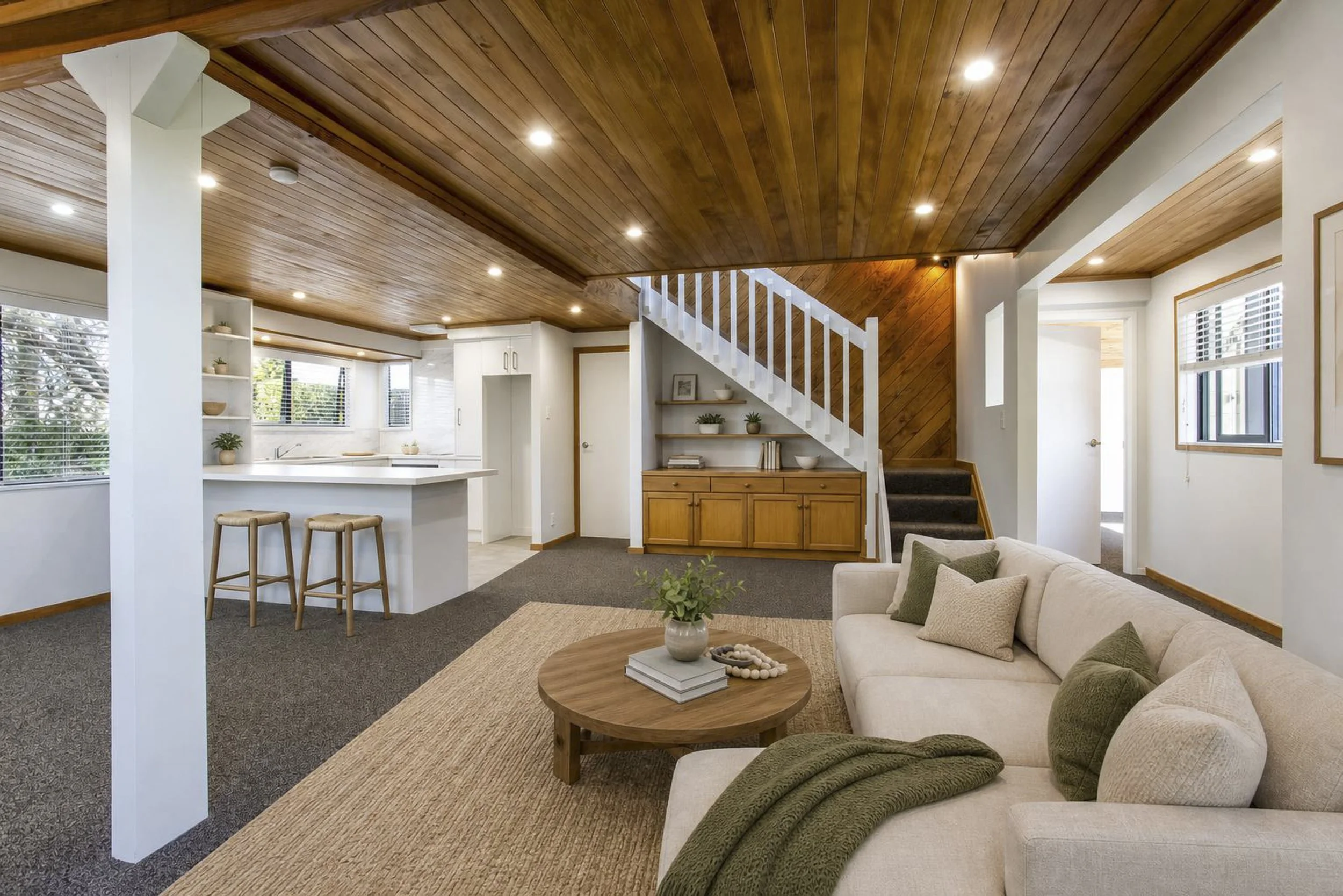 Living room with beige couch, green pillows, wooden coffee table with books and decorative items, beige area rug, window with white blinds, staircase with wooden steps and white railing, white kitchen cabinets with window, wood ceiling with recessed lighting.