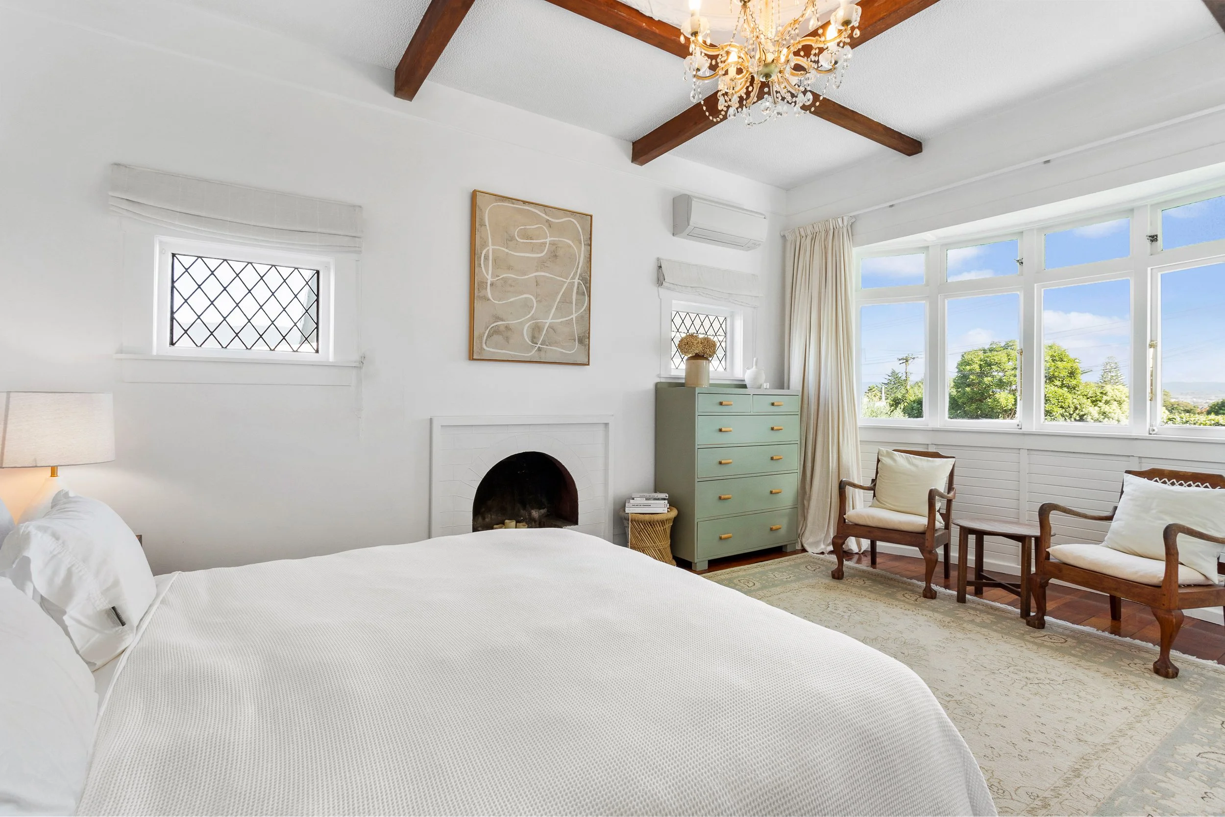 A cozy bedroom with a white bed, a green dresser, wooden chairs, and large windows showing a view of trees and blue sky, decorated with a chandelier and artwork on the wall.