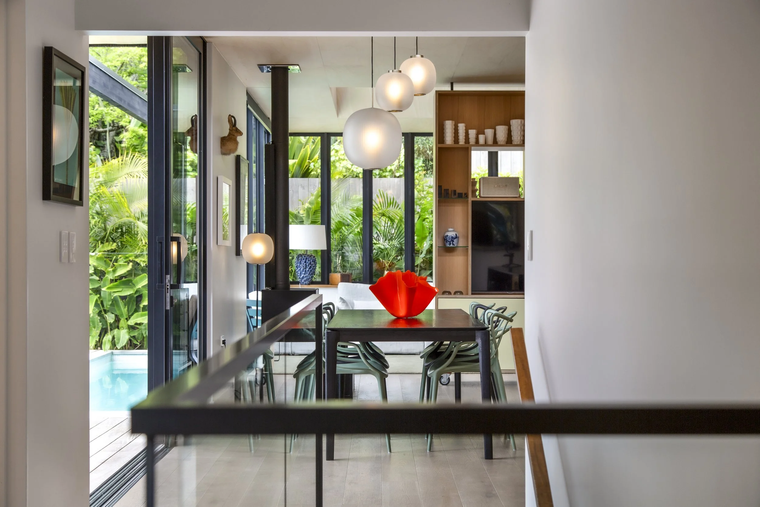 A modern dining room with a large dark wooden table and teal chairs, decorated with a red vase, with sliding glass doors opening to a lush green outdoor area with trees and plants, and white pendant lights hanging from the ceiling.
