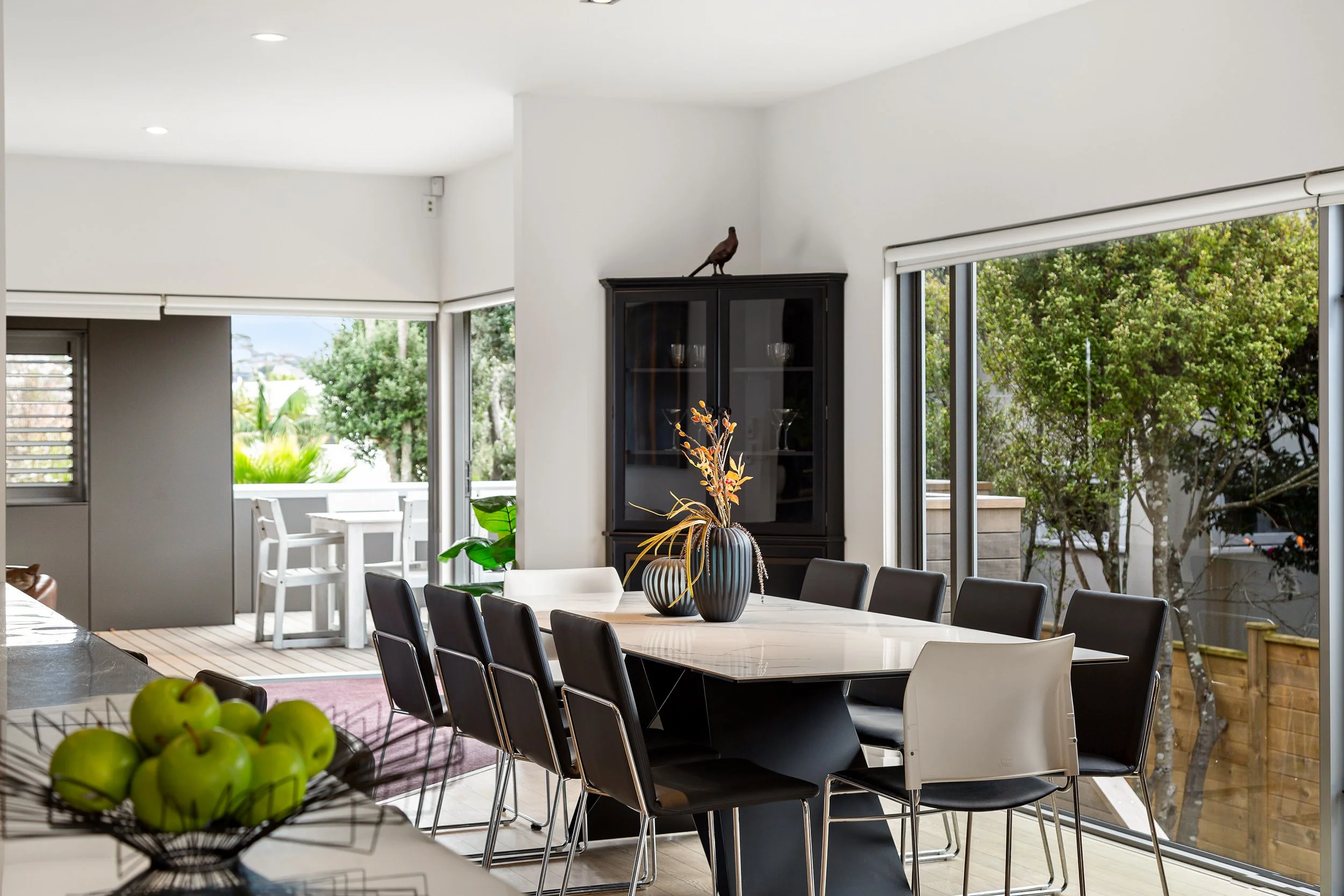Modern dining room with a large table surrounded by black and white chairs, a decorative vase with plants, and large windows showing outdoor greenery.