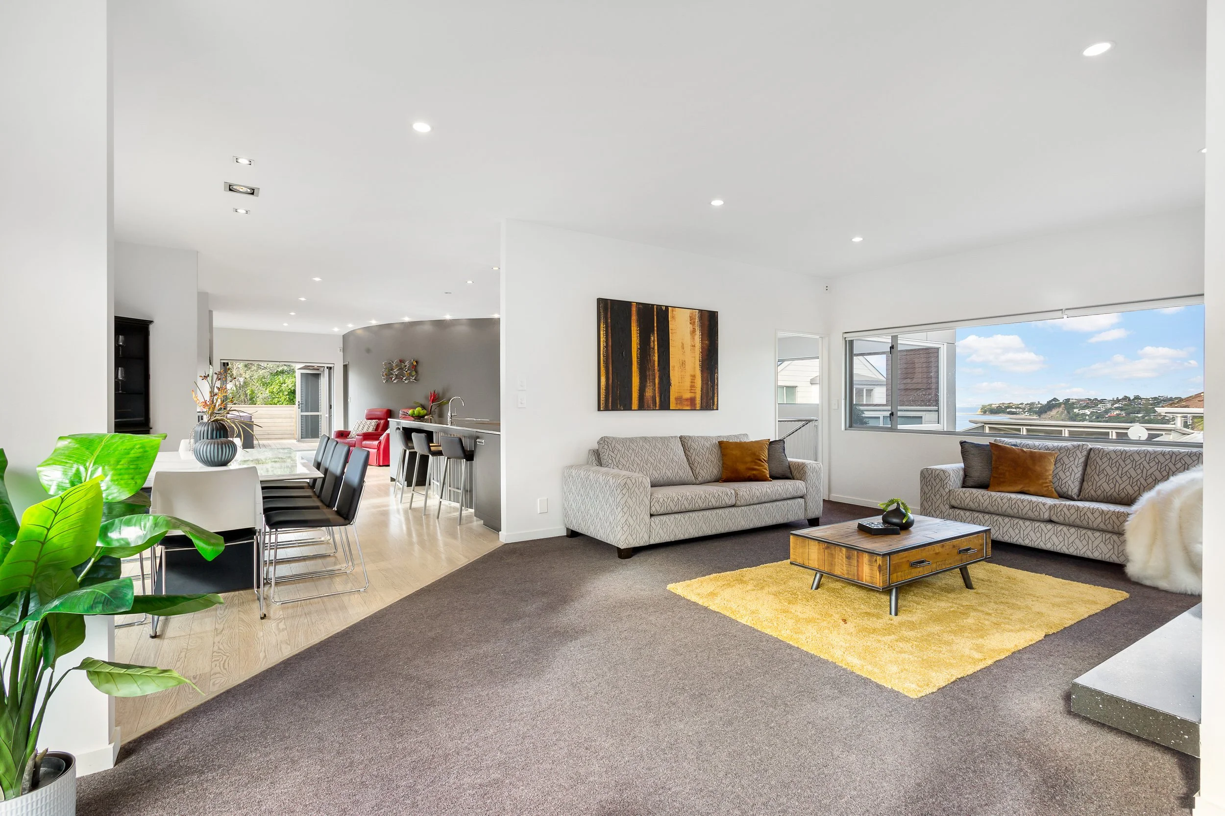 Open-concept living and dining area with gray sofas, a wooden coffee table, a yellow rug, large windows showing a view of a neighborhood, and a dining table with black chairs, decorated with vases and plants.