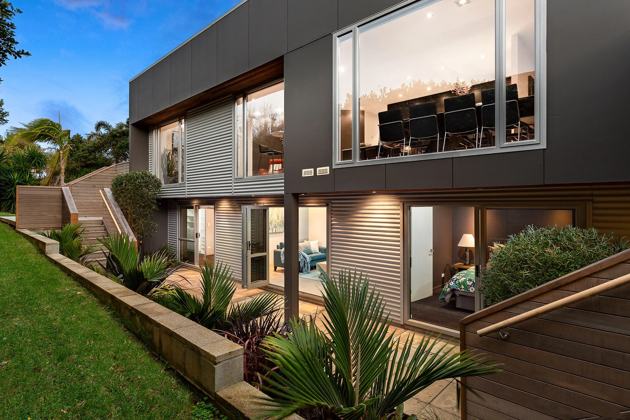 Modern multi-story house with large glass windows, outdoor patio, lush green lawn, and garden, illuminated interior in the evening.