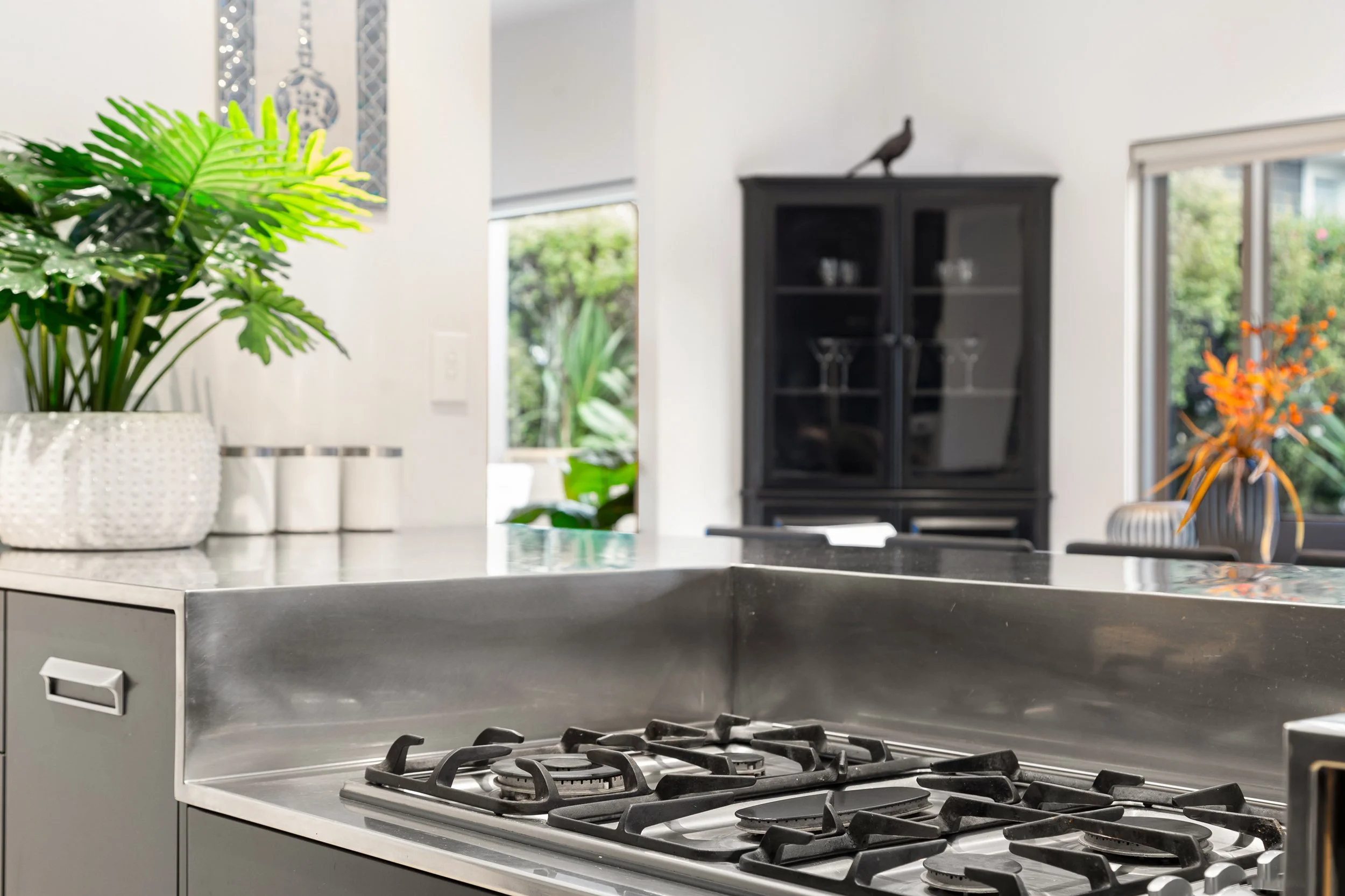 A modern kitchen with a black stove and a gray countertop, decorated with a green potted plant, visible windows, and a black cabinet with a bird sculpture on top.