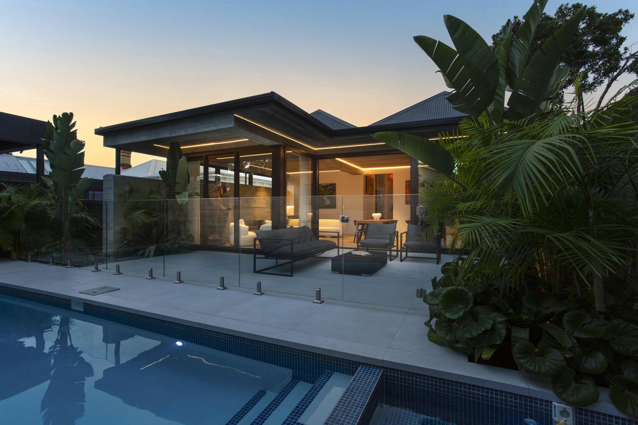 Modern house with large glass windows and a lit patio, surrounded by lush tropical plants, at dusk. A swimming pool is visible in the foreground.