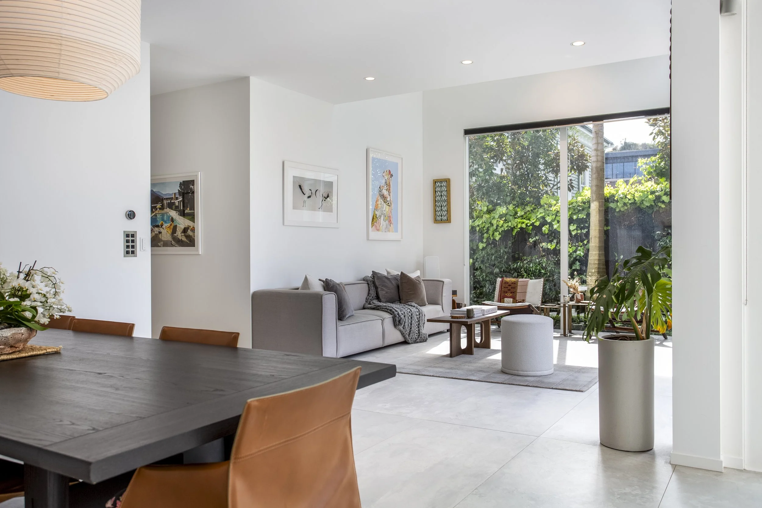 Modern living room with white walls, large window showing greenery outside, framed artwork on walls, beige sofa with pillows, small coffee table, decorative plants, and recessed ceiling lights.