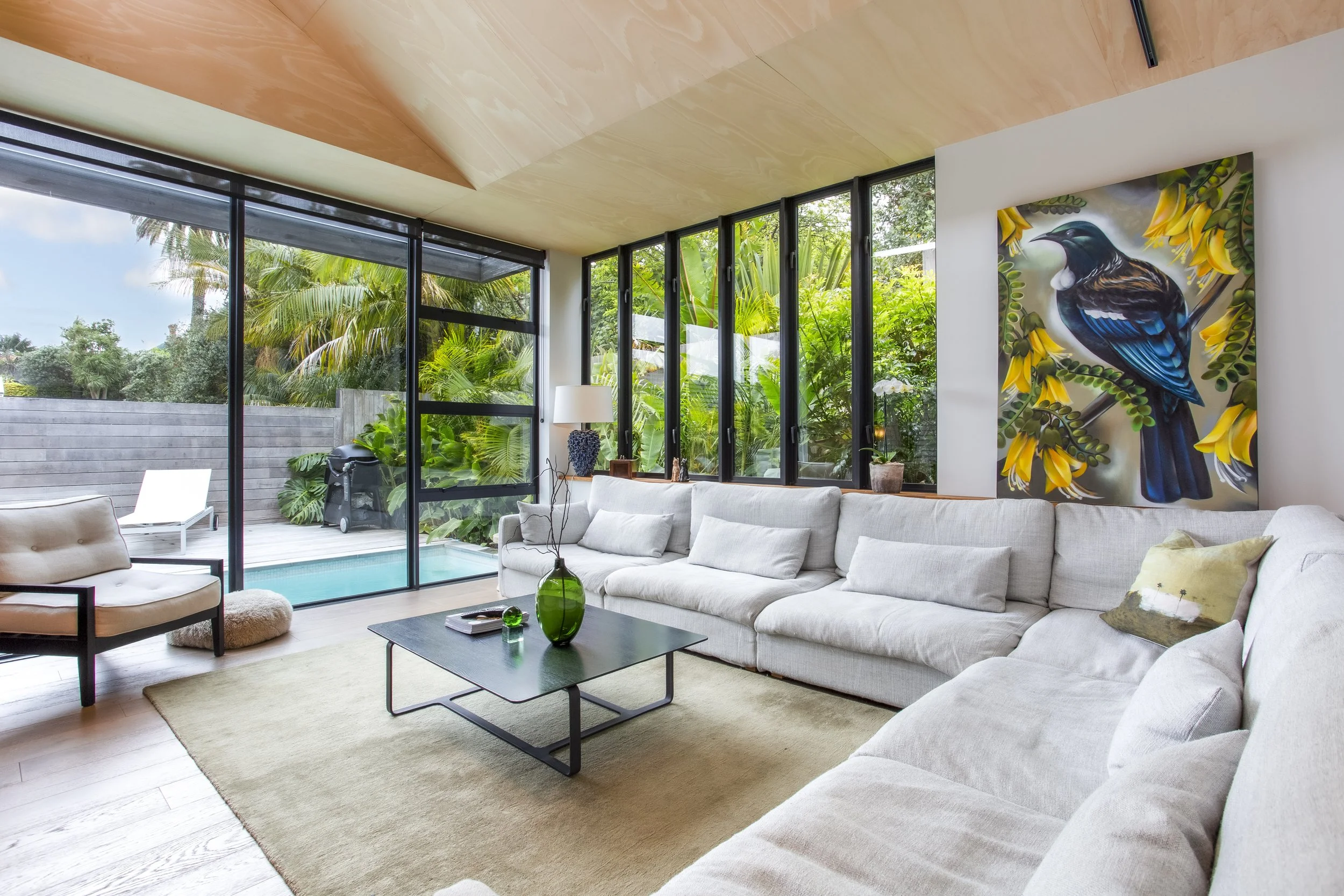 Living room with large windows showing outside greenery, a white sectional sofa, a piece of colorful bird artwork on the wall, a black coffee table with a green vase, and a beige rug.