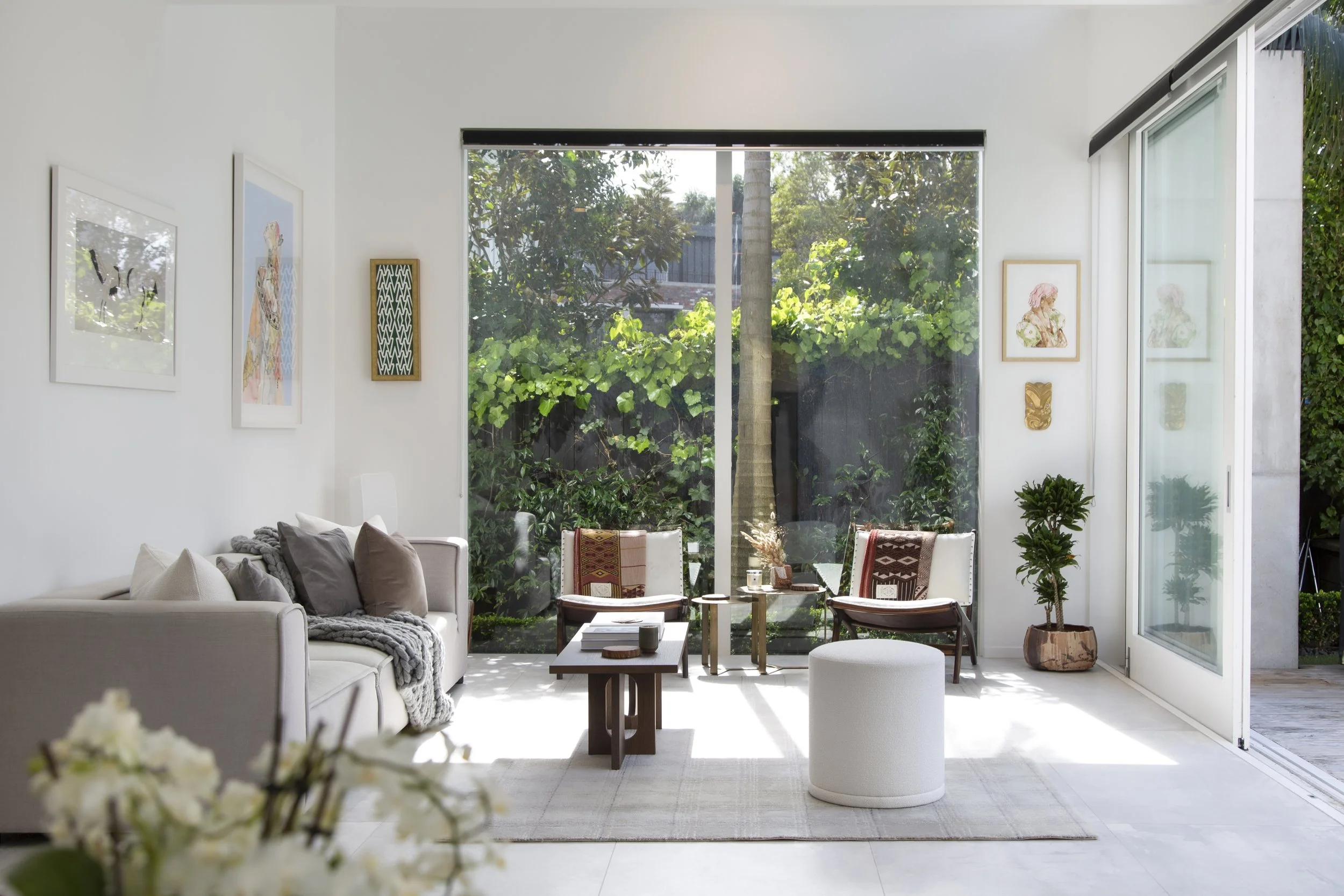 Bright living room with large window and patio door, white walls, framed artwork, white sofa with cushions, wooden coffee table, chairs, indoor plants, and outdoor greenery visible through window.