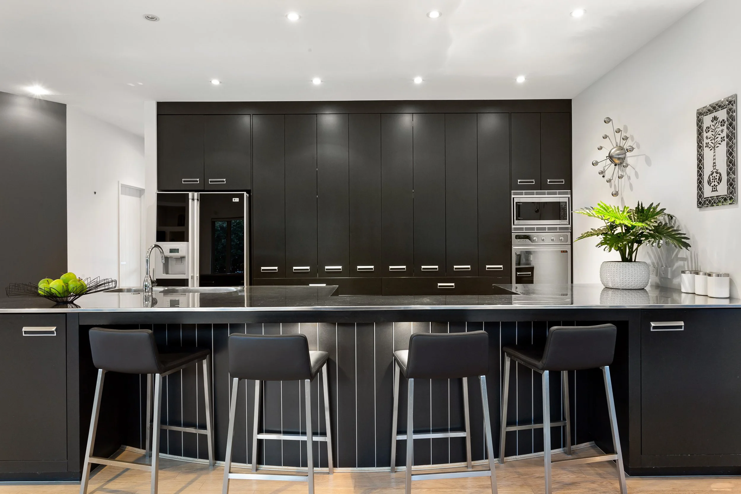Modern kitchen with black cabinets, stainless steel appliances, a large marble island with a sink, black chairs, a potted plant, and decorative wall art.
