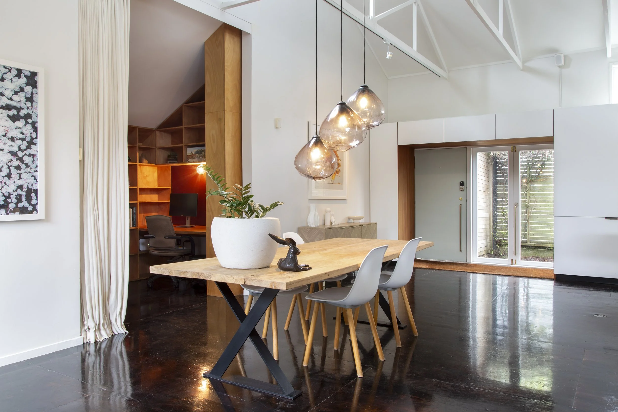 A modern dining room with a wooden dining table, white chairs, and a large pendant light fixture. The ceiling is vaulted with white beams, and there is a sliding glass door leading outside. A potted plant and decorative items are on a sideboard. An open kitchen or office space with wooden shelving and a desk is visible in the background.