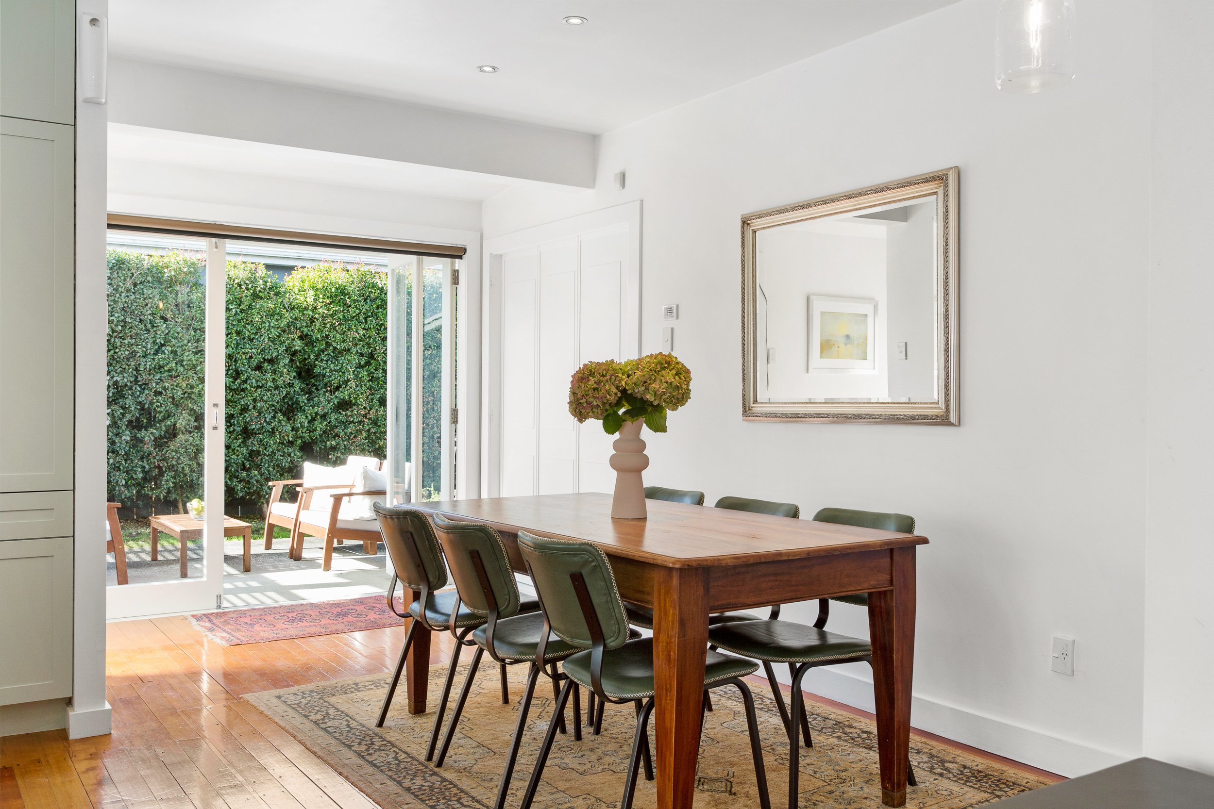 Interior of a dining area with a wooden table, six chairs, a vase with flowers, a mirror on the wall, and an open sliding glass door leading to a backyard with outdoor furniture and greenery.
