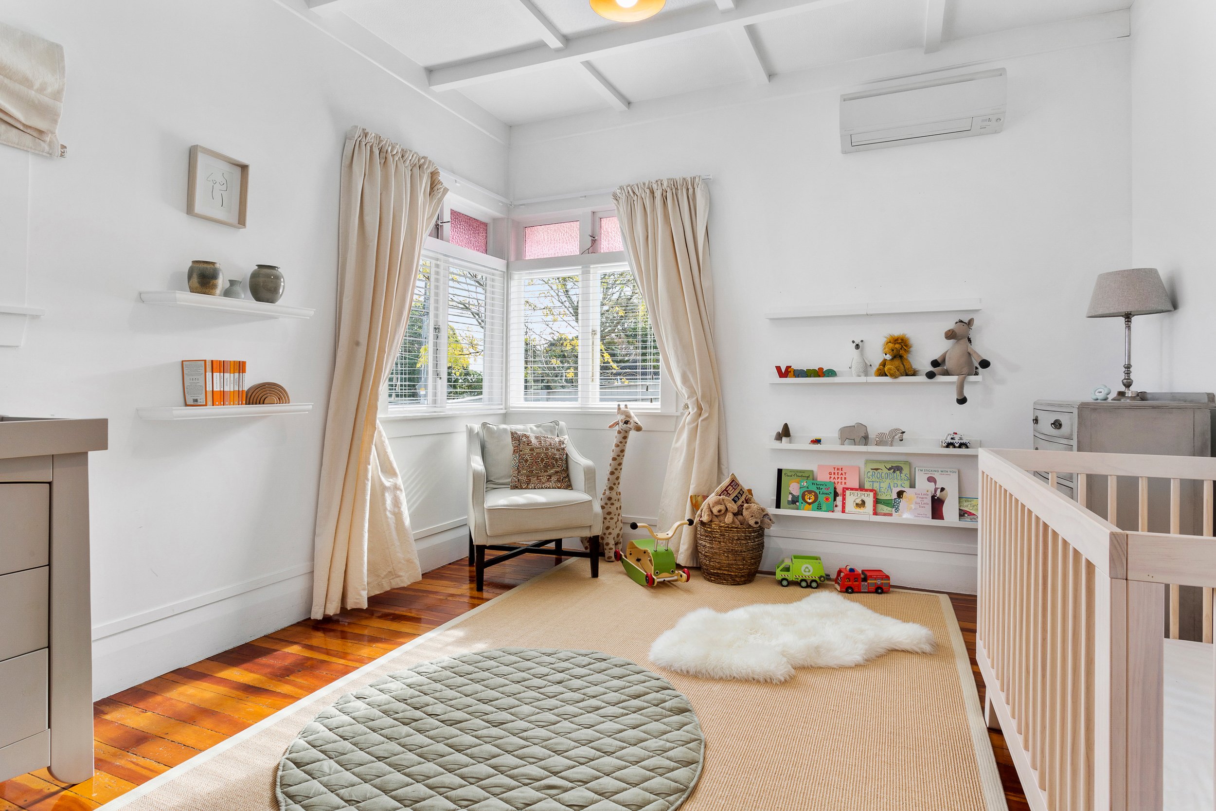 A bright children's bedroom with white walls and a large bay window with cream curtains. The room has a white shelf with toys, books, and plush animals, including a giraffe, a lion, a sheep, and an elephant. There is a white armchair with a patterned