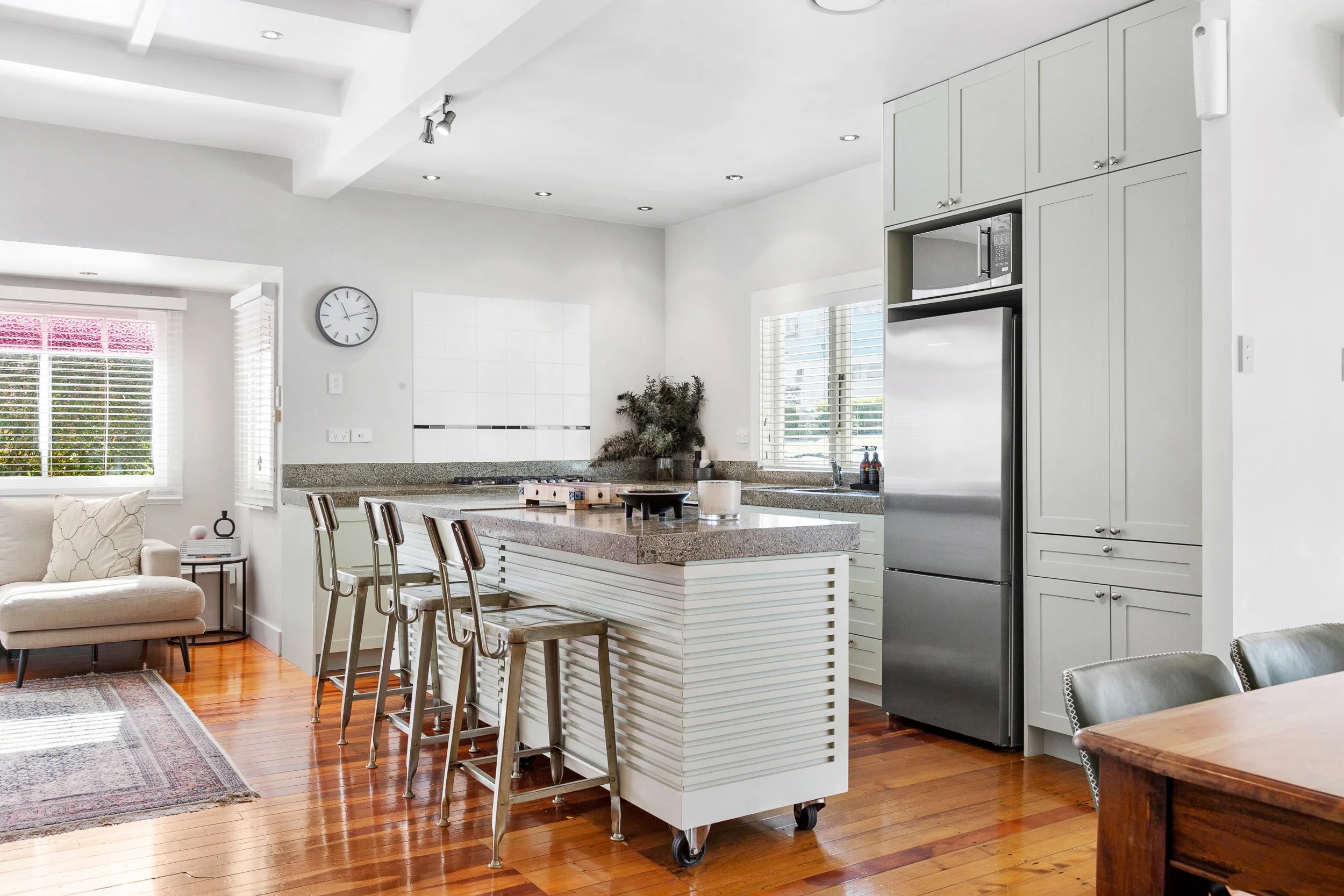 A modern, white kitchen with a large island, stainless steel refrigerator, microwave, white cabinets, and a window with blinds. There is a living room area with a beige sofa and a hardwood floor.