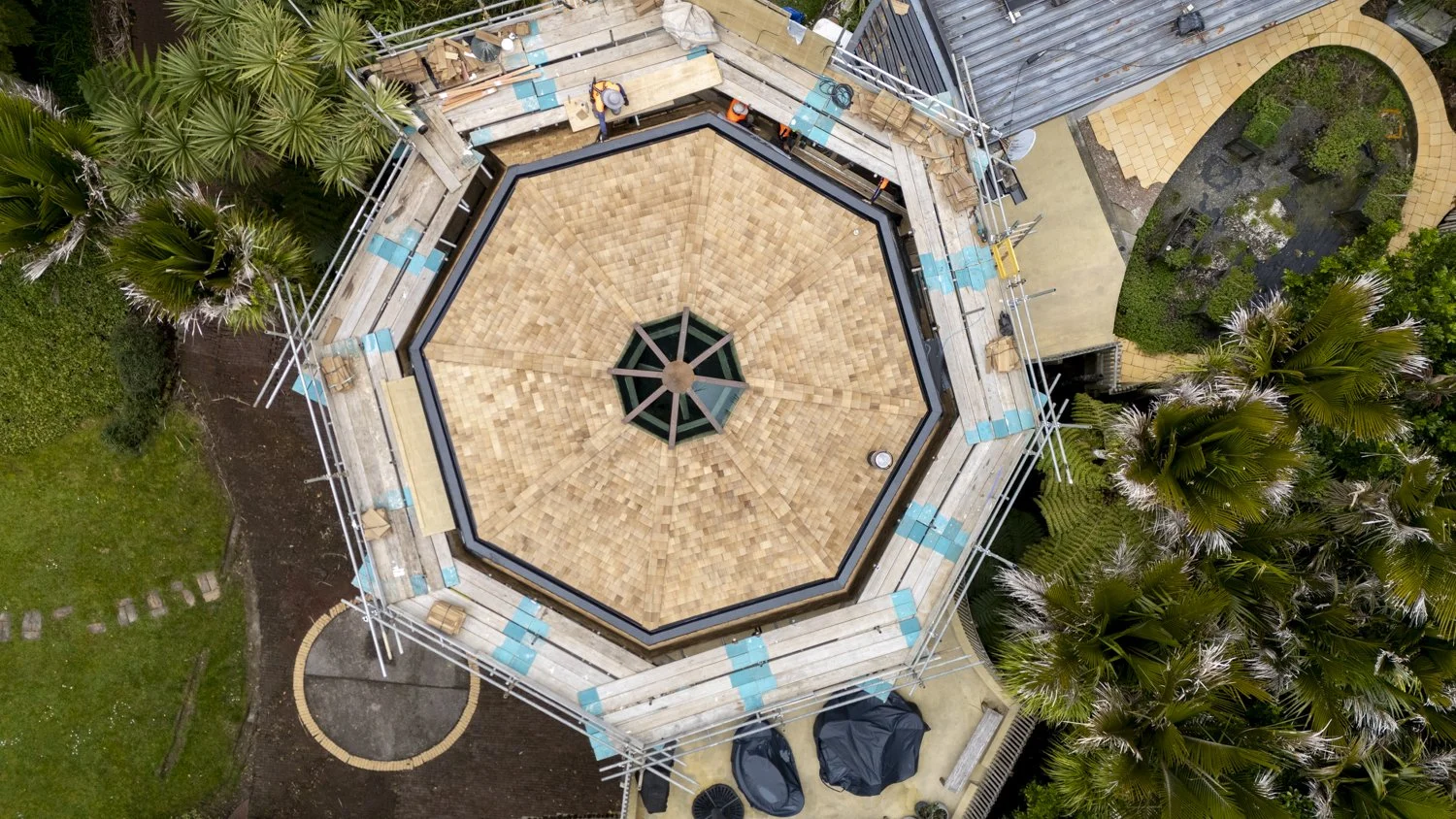 An aerial view of a building under construction with a hexagonal wooden roof. Surrounding the building are scaffolding, construction materials, and nearby palm trees.
