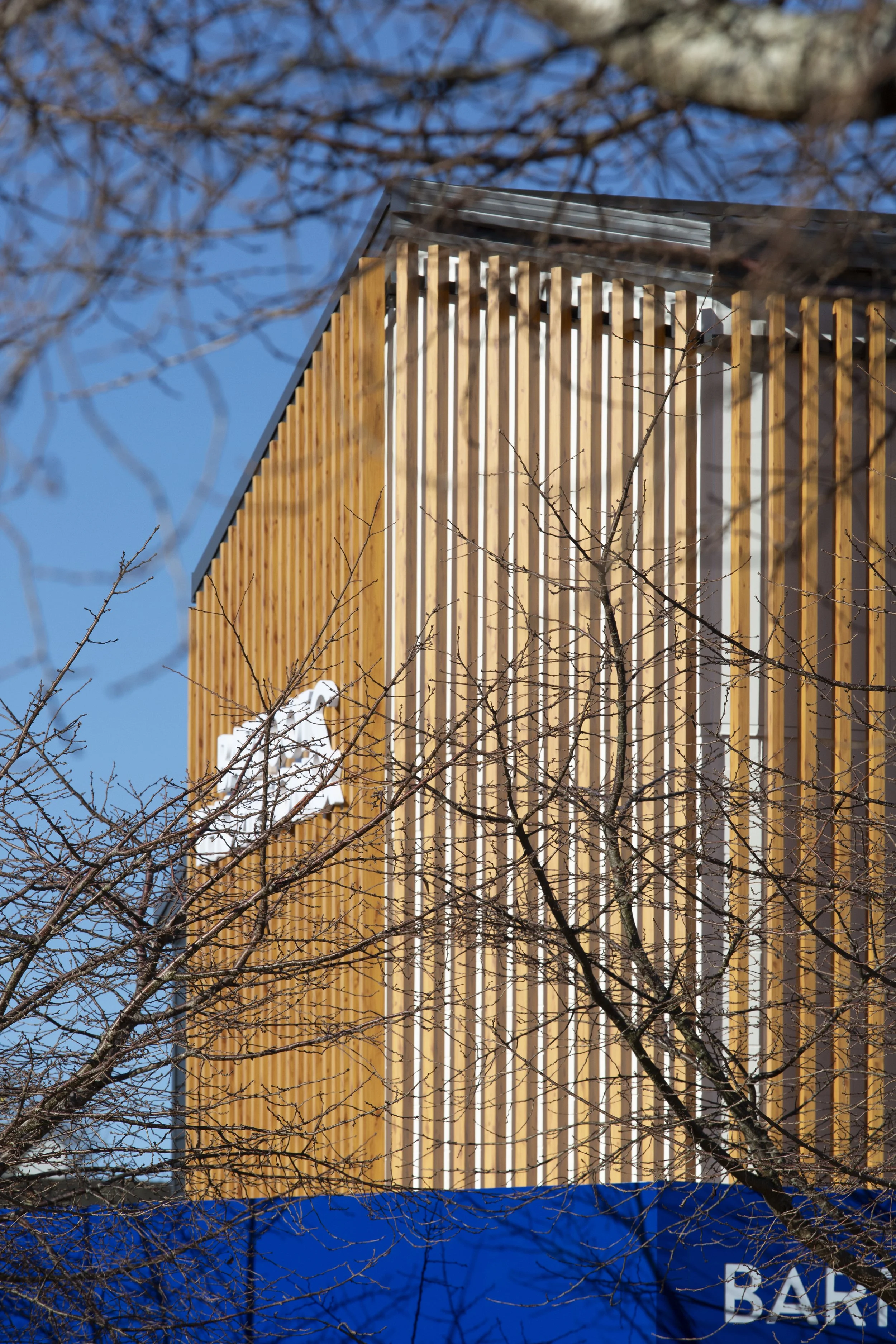 Tall building with vertical wooden slats and a metallic roof, partially obscured by leafless tree branches, against a clear blue sky.