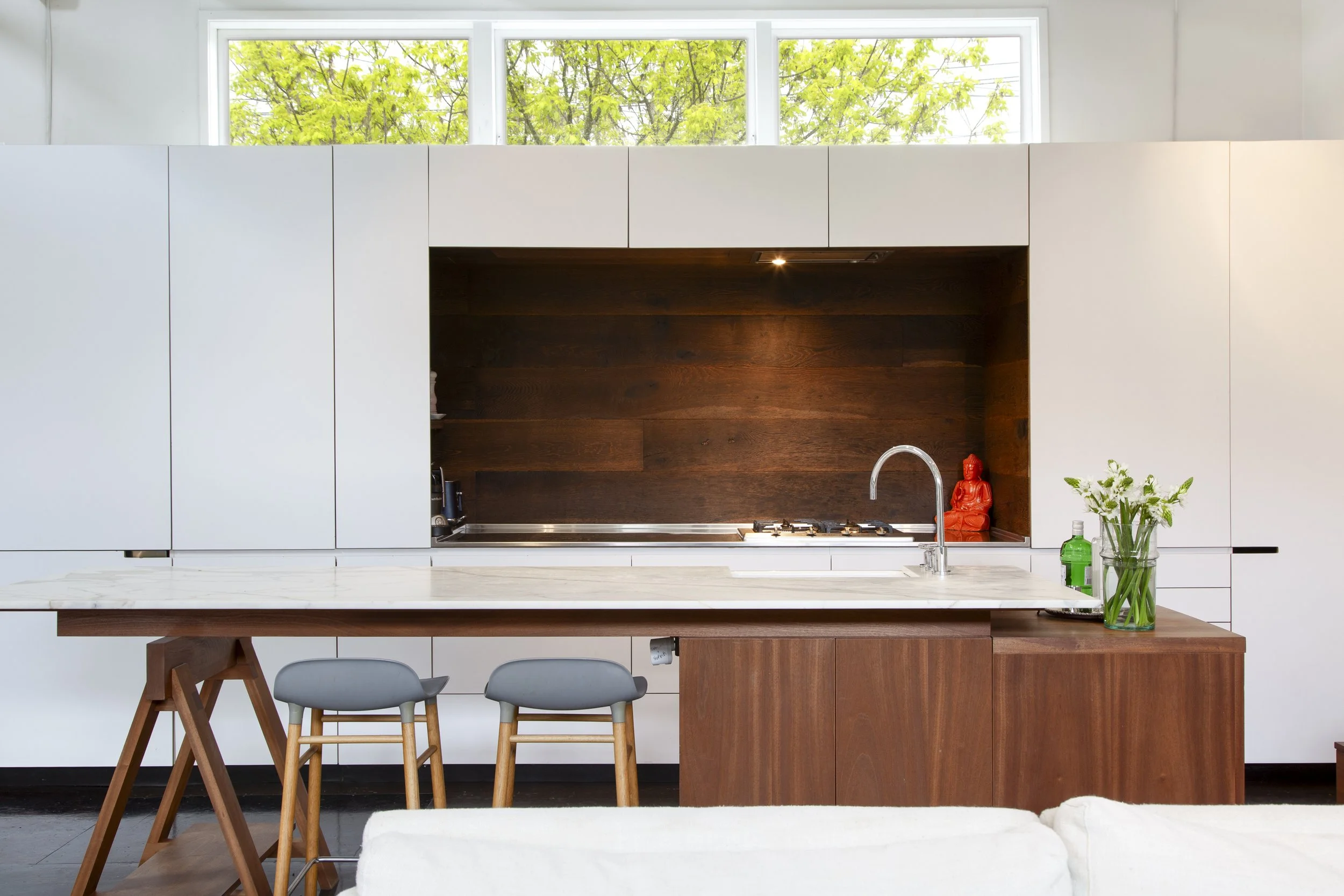 Modern kitchen with white cabinets, a dark wood backsplash, and a window with green trees outside. There is a kitchen counter with a sink, a small orange Buddha statue, a green liquid soap bottle, and a vase with white flowers.