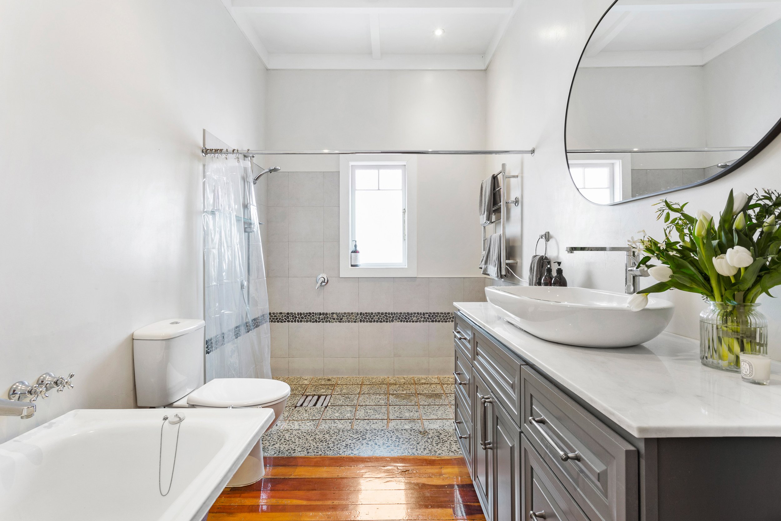 Bright, modern bathroom with white walls, a bathtub, a toilet, a vanity with a sink, and a large mirror. Decor includes fresh flowers in a vase, towels, and natural light from a window.