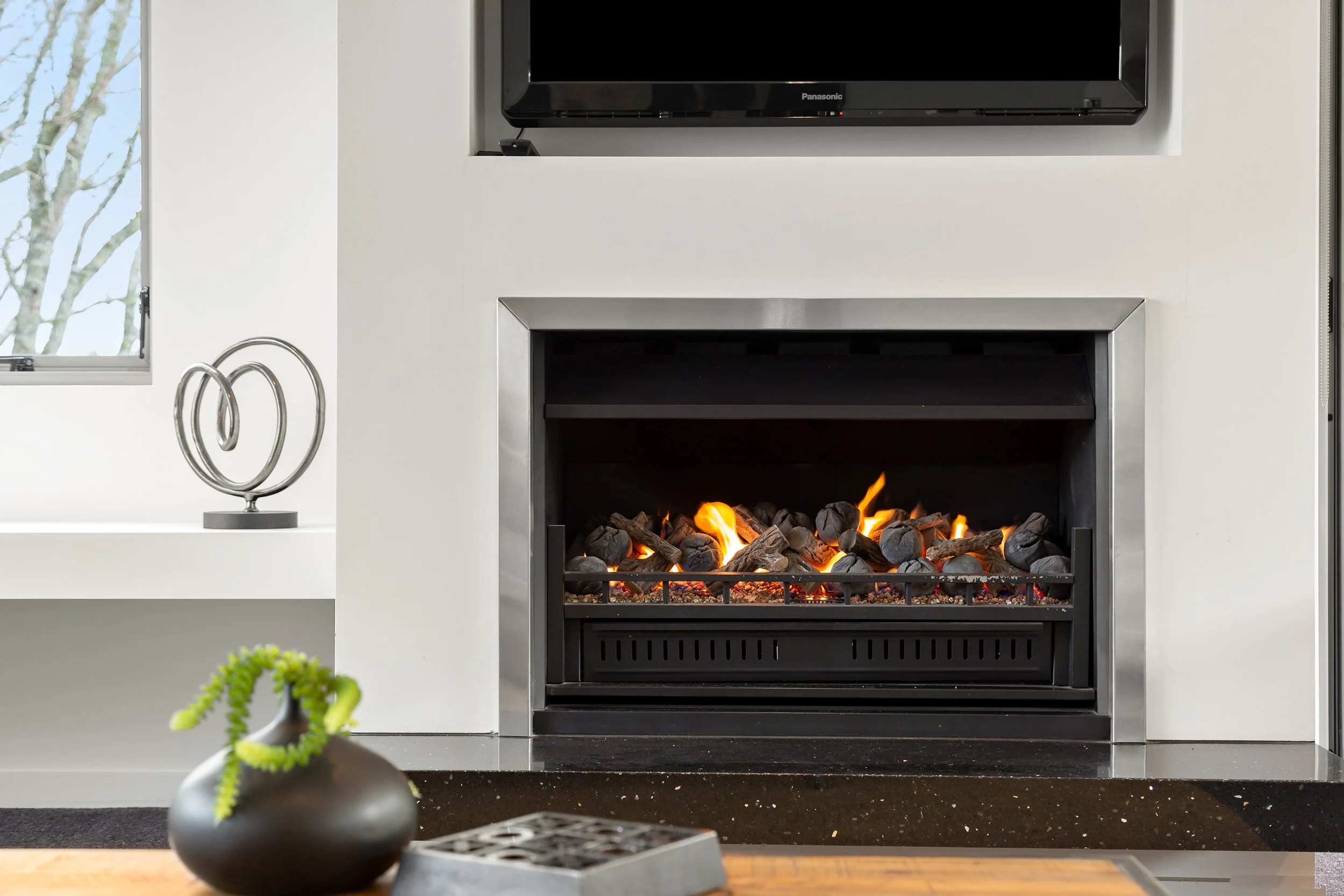 A modern living room with a wall-mounted flat-screen TV above a fireplace with glowing logs and flames, a window on the left showing trees outside, and a decorative metal sculpture on the white shelf on the left.