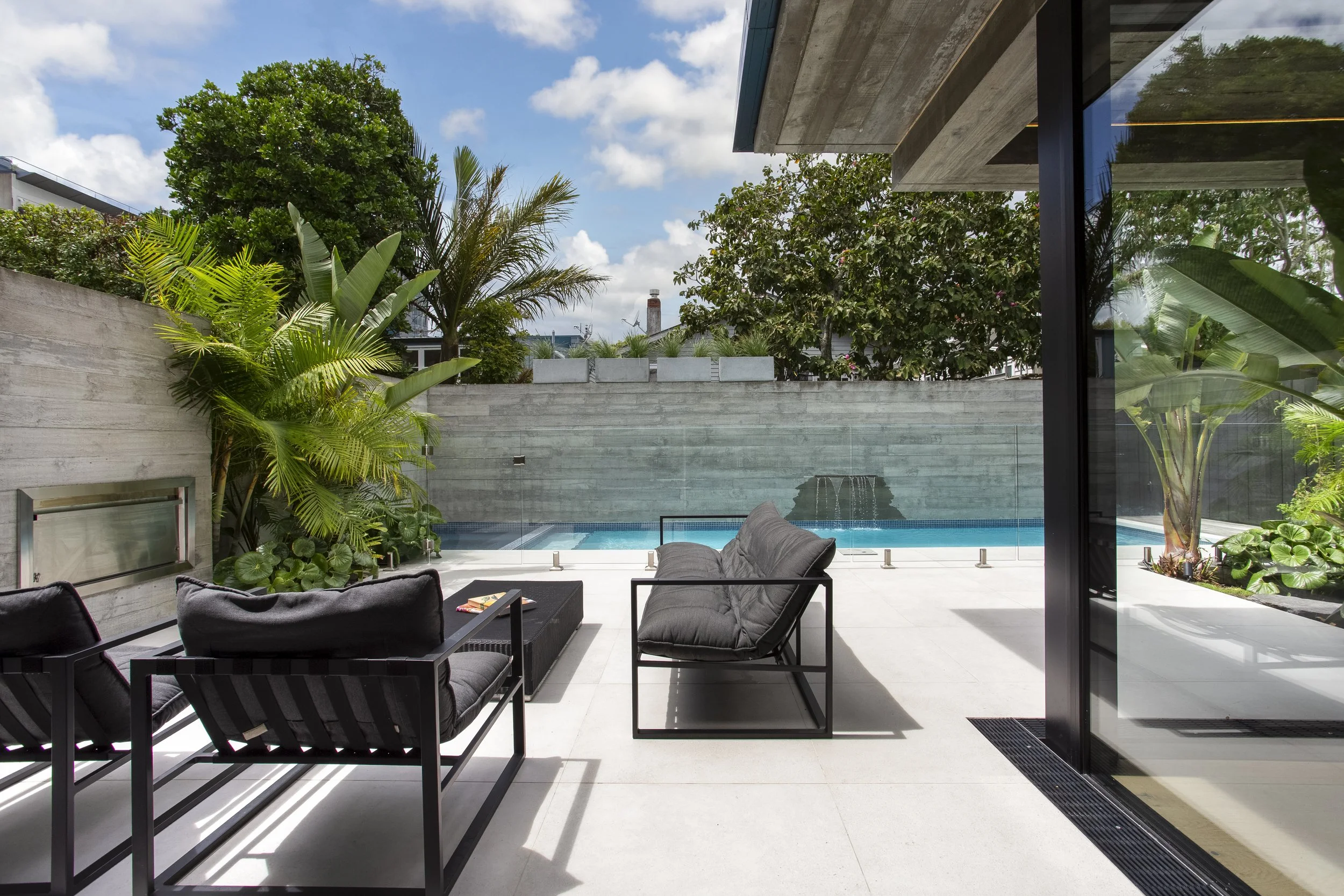 Outdoor patio area with black cushioned chairs and a sofa, overlooking a backyard with a swimming pool, surrounded by a concrete wall and lush green trees and plants, with a partly cloudy sky overhead.