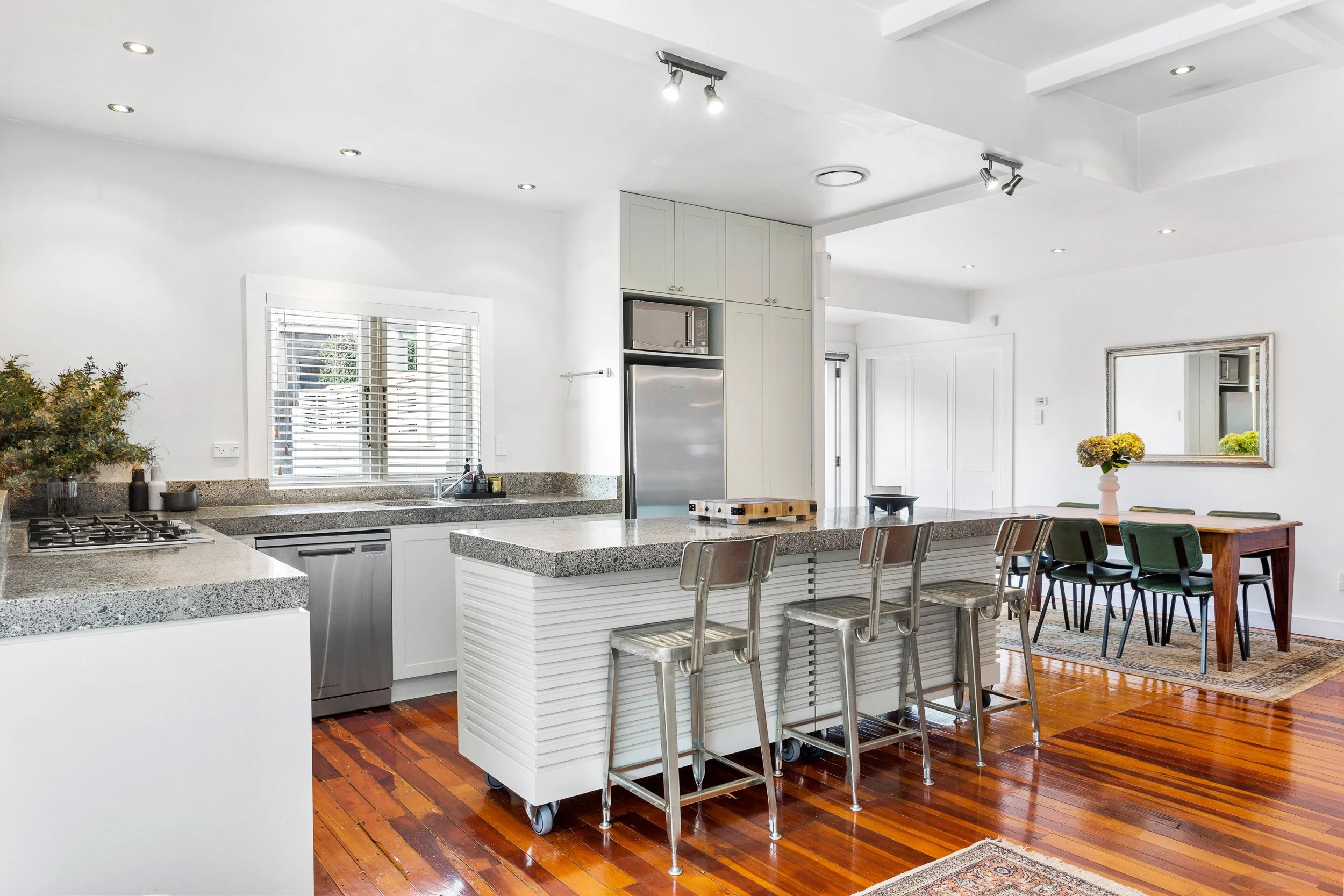 A modern kitchen with white cabinets, gray countertops, a kitchen island with metal stools, and hardwood floors. There are large windows with blinds, a stainless steel refrigerator, microwave, and a dining table with chairs and a vase of flowers.