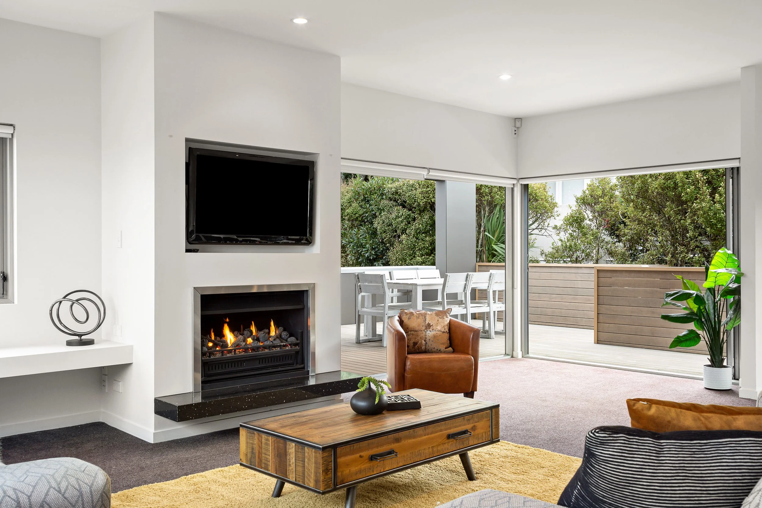 Interior view of a modern living room with a fireplace, brown leather chair, wooden coffee table, and a large window opening to a patio with outdoor furniture and lush greenery.