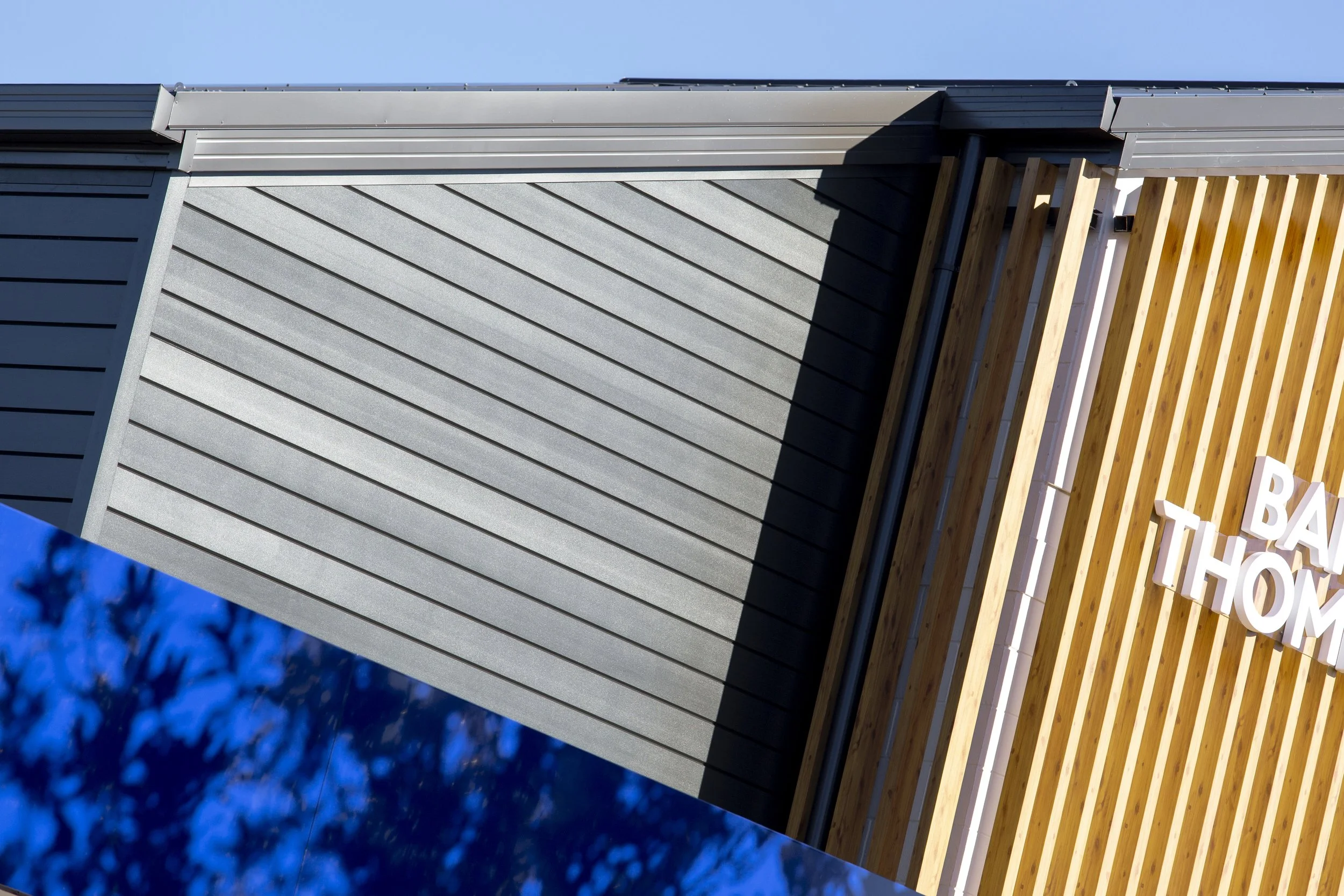 Close-up of a modern building's exterior wall with gray horizontal siding, wooden slats on the right side, and a portion of a sign with white letters, against a blue sky background.
