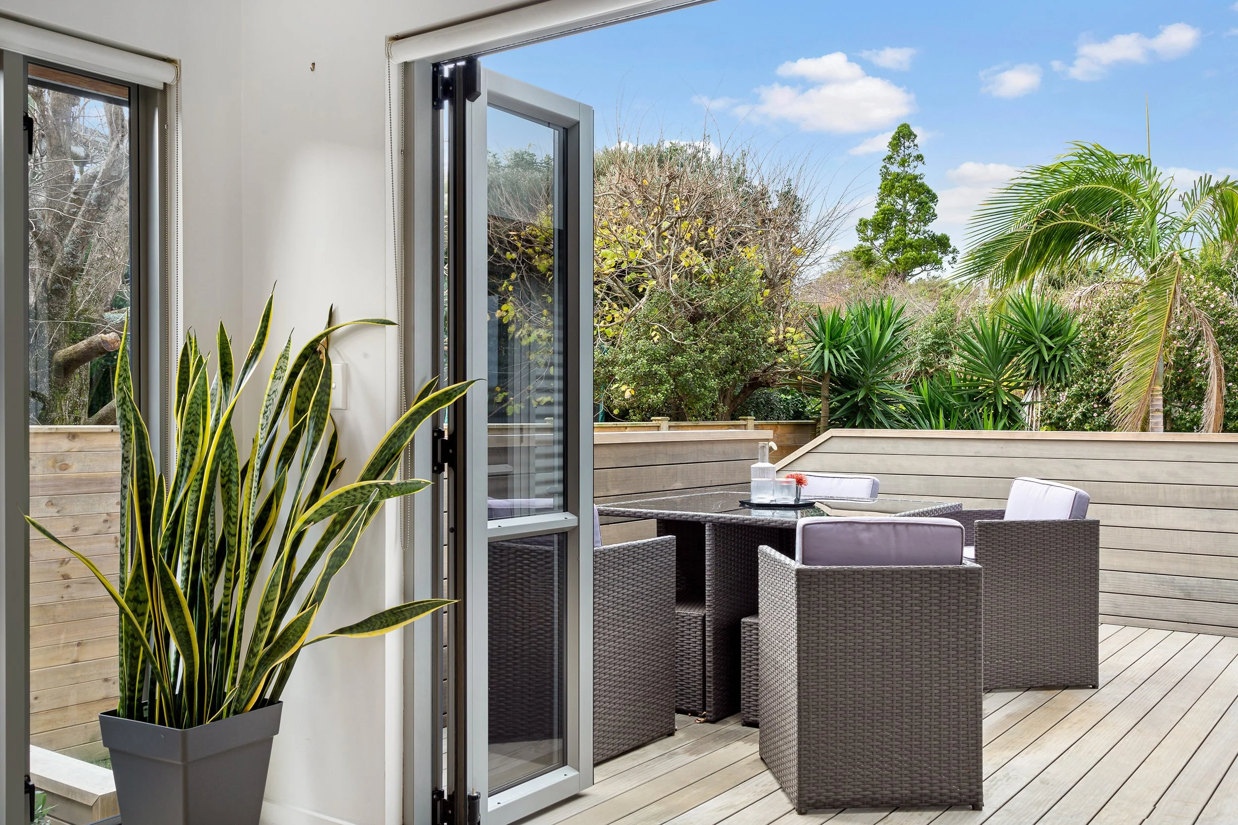 View of a patio with outdoor wicker chairs around a glass table, surrounded by a wooden fence and lush green trees and plants, seen through open sliding glass doors from inside a room with a potted plant in the foreground.
