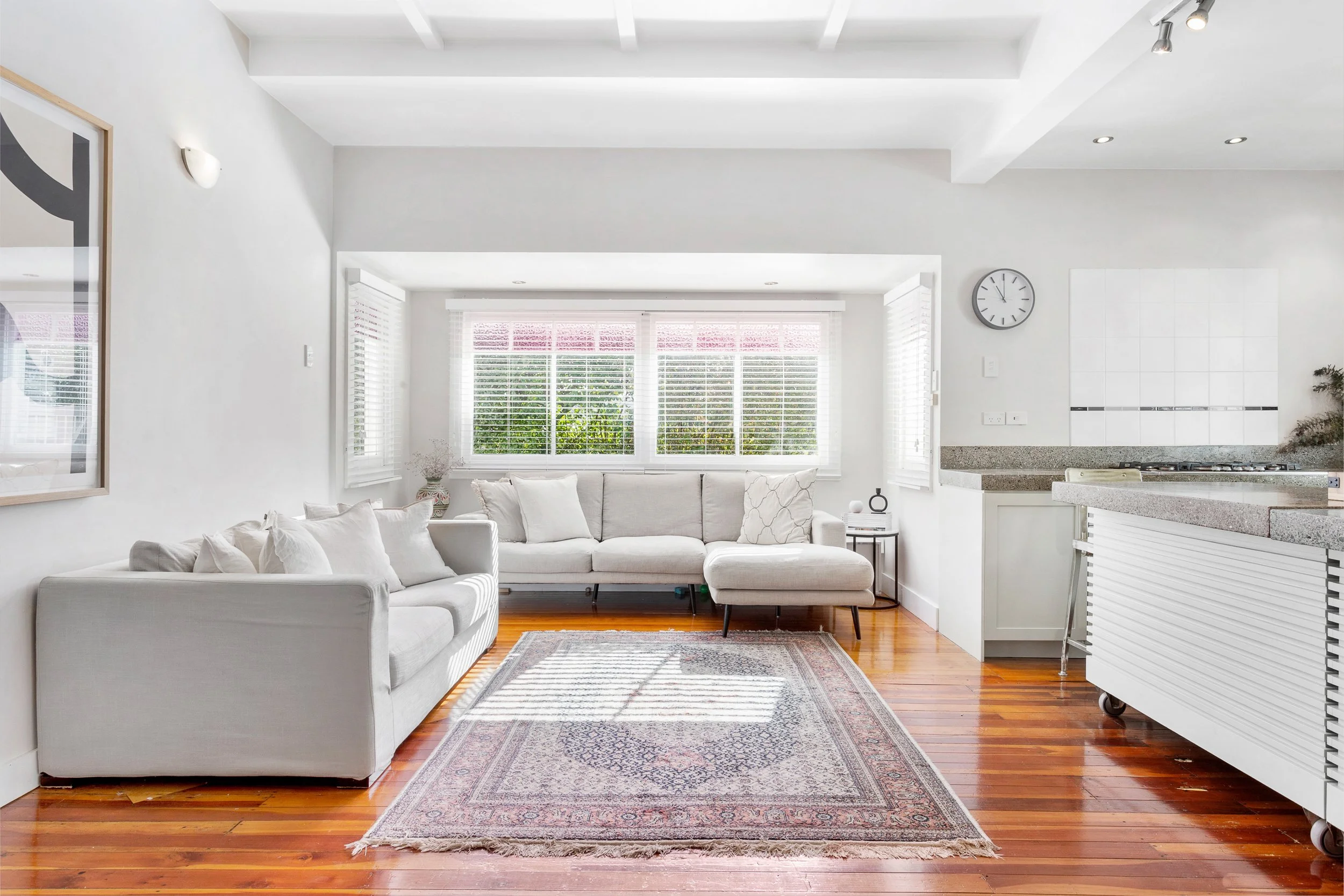 A bright, modern living room with white walls, a white sectional sofa with multiple pillows, and a large window with white blinds. There is a patterned area rug on hardwood floors, a wall clock, and a partial view of a kitchen with granite countertop