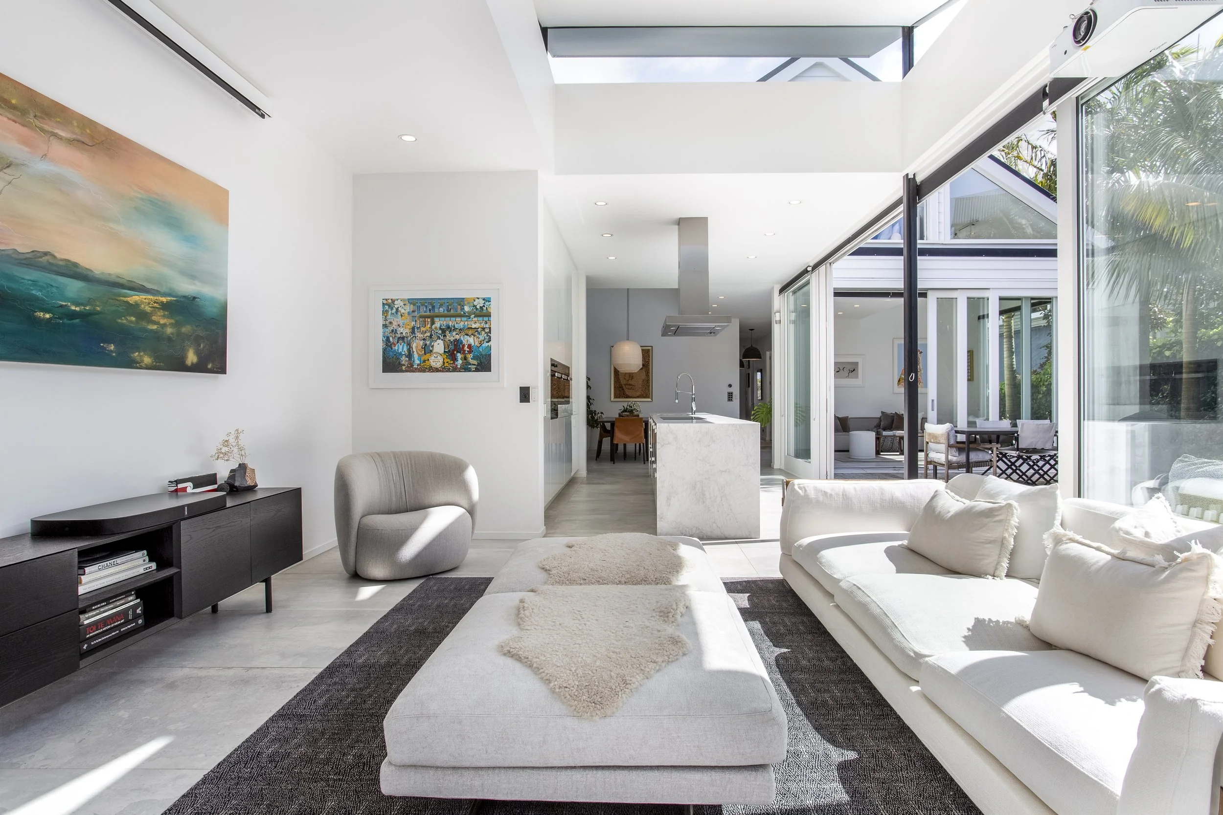 Modern open-plan living room with white walls, a large white sofa with plush throws, a black media console with books, and artwork on the walls. Large glass sliding doors open to an outdoor patio with greenery, and natural light floods the space through a skylight.