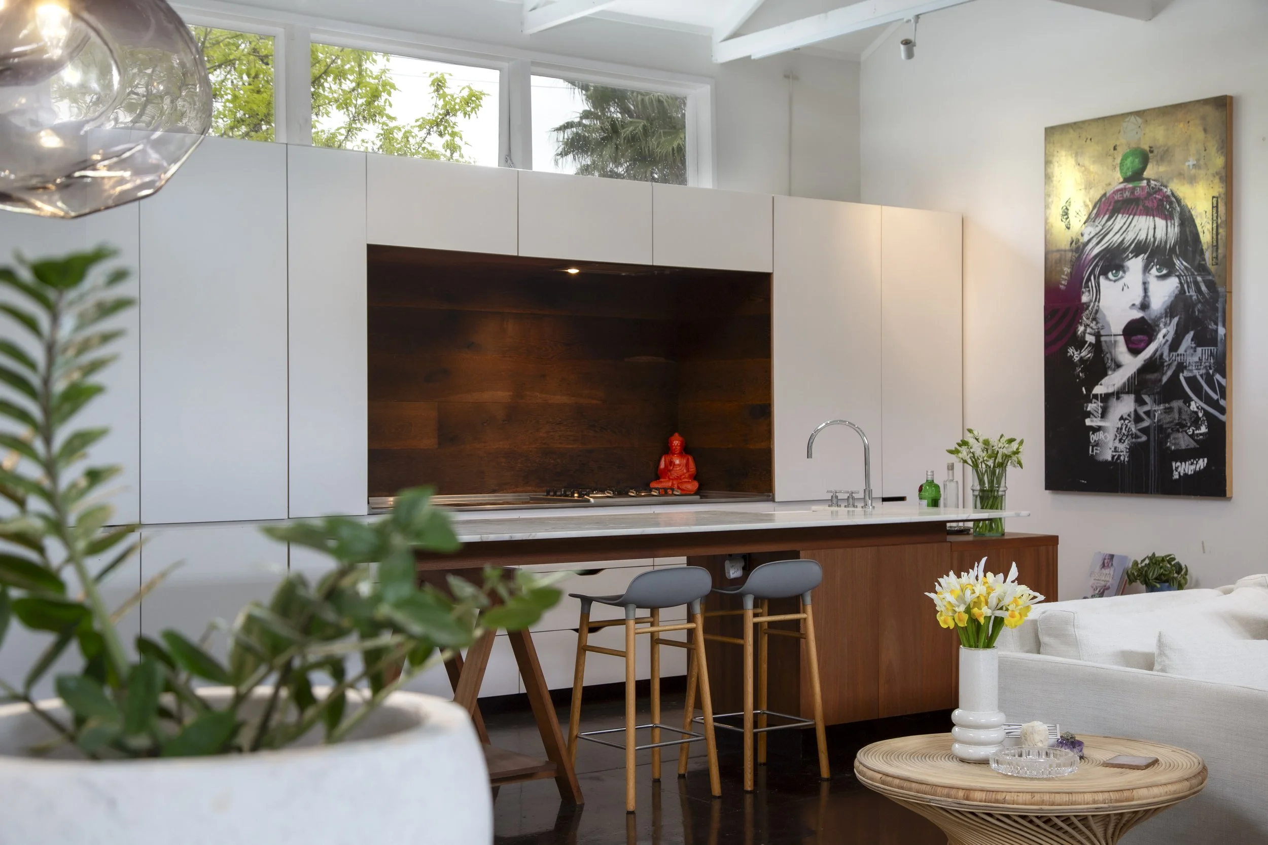 Modern kitchen with white cabinets, wooden accents, and colorful artwork of a woman with a pink headband on the wall. Green plants and flowers decorate the space, which is illuminated by natural light from large windows.