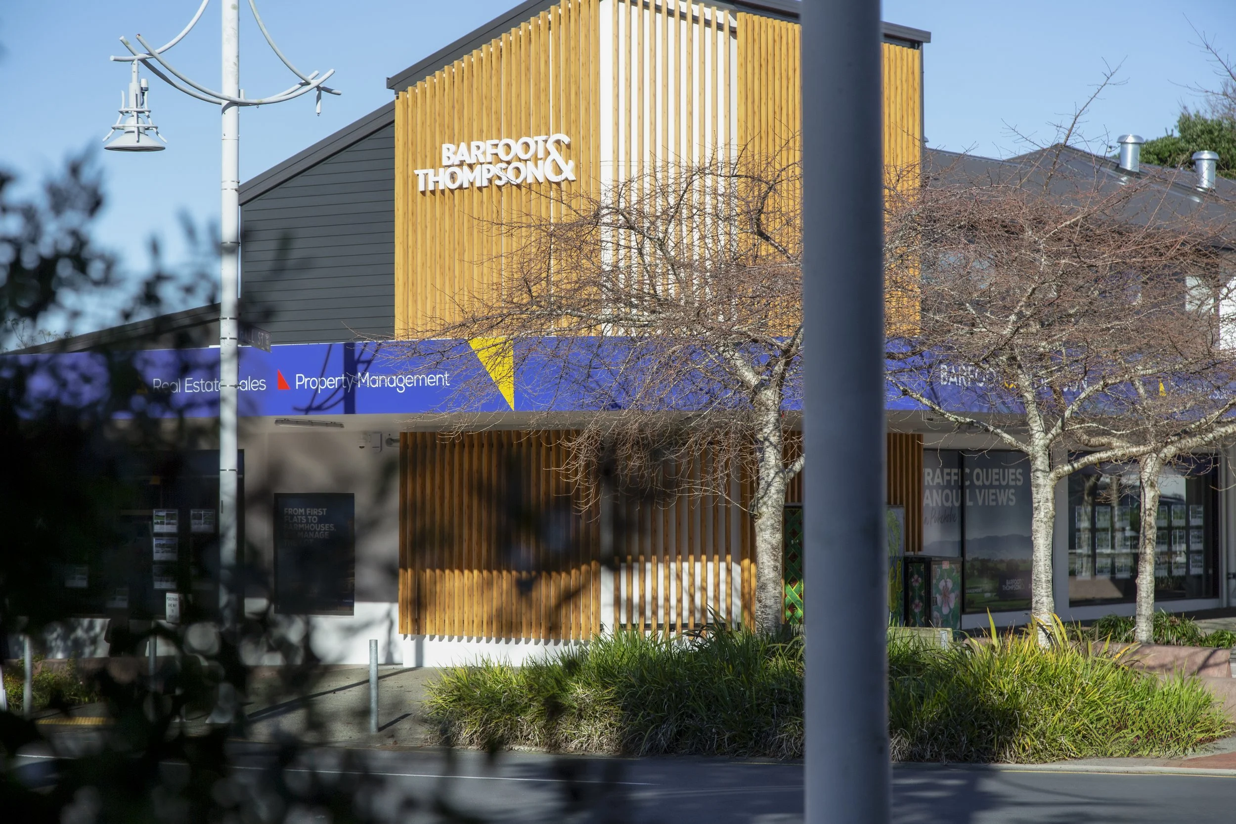 A modern building with a sign that reads 'Barfoot & Thompson' in white letters on a wooden panel. The building is surrounded by leafless trees and a traffic light pole, with a blue sky overhead.