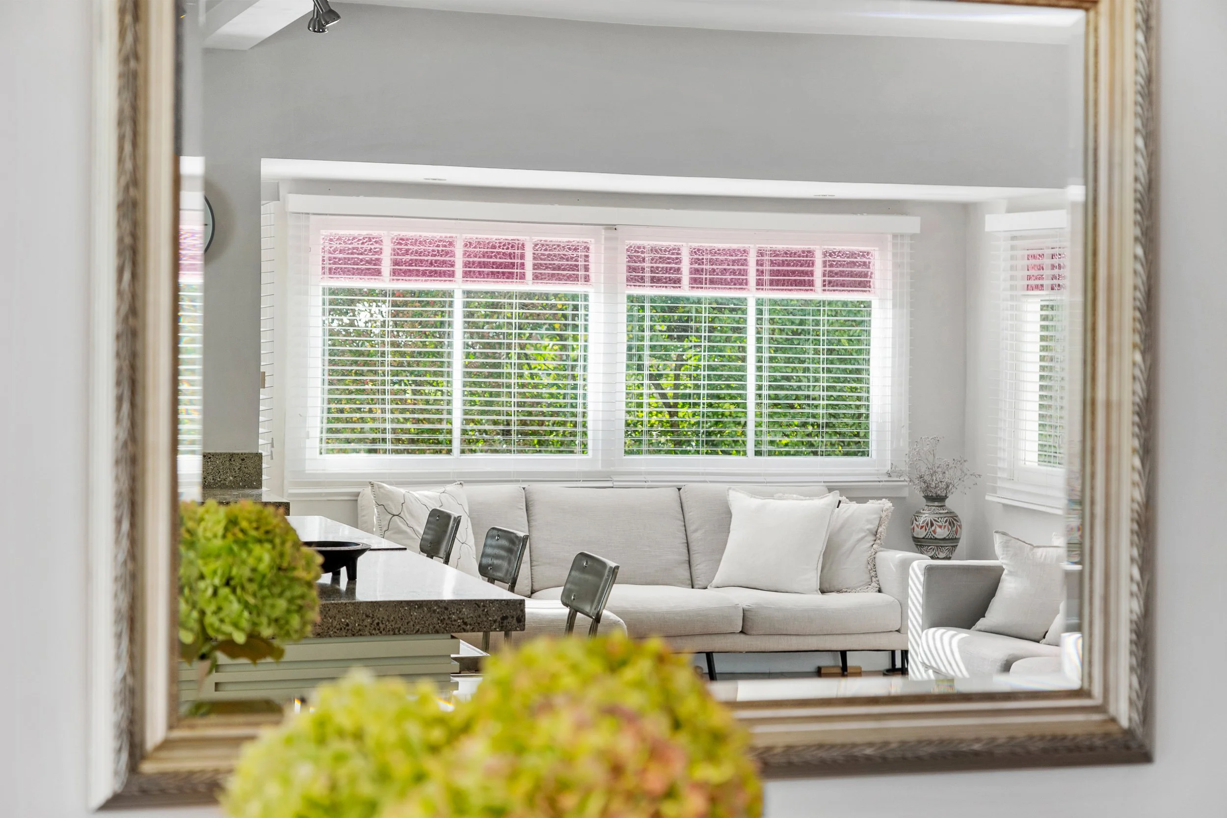 A living room with a large window, white blinds, a beige sofa with white pillows, a round table with chairs, and decorative accents, viewed through a mirror on a wall.