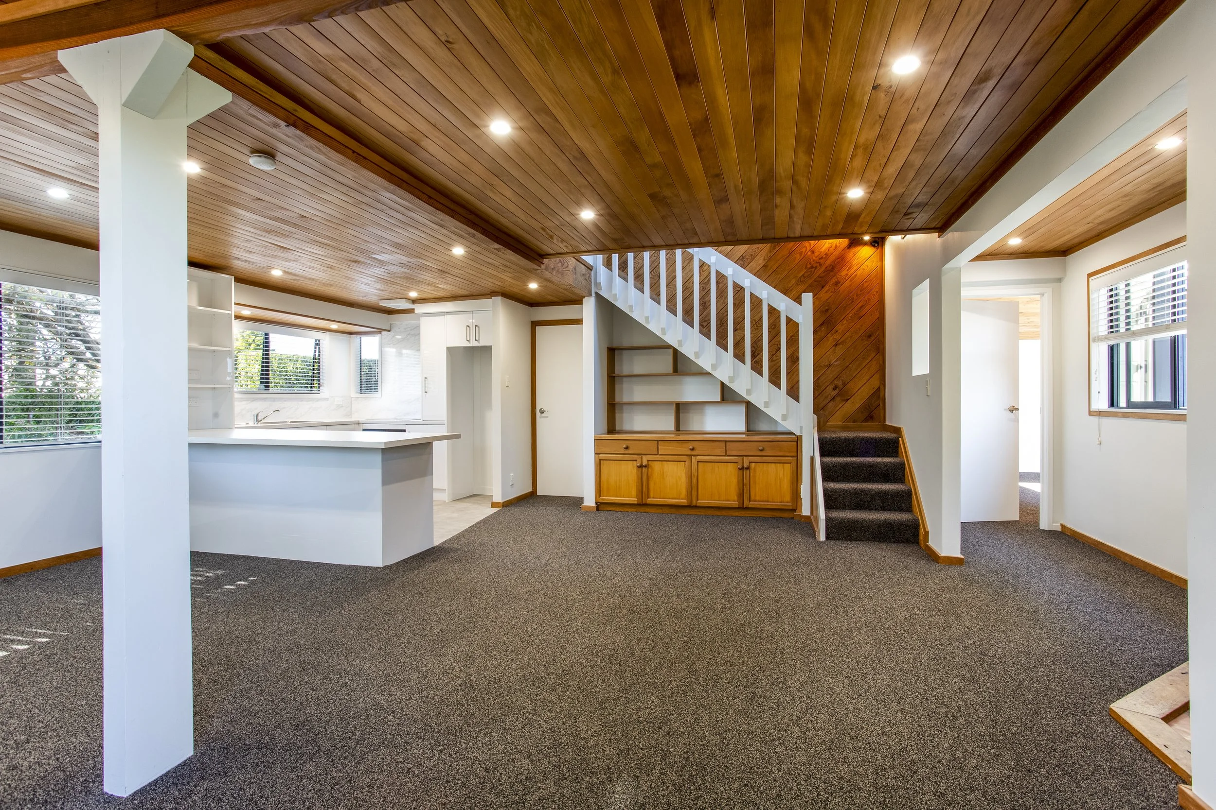 Living room with wooden ceiling and staircase, large windows with blinds, white walls, carpeted floor, and kitchen area in the background.