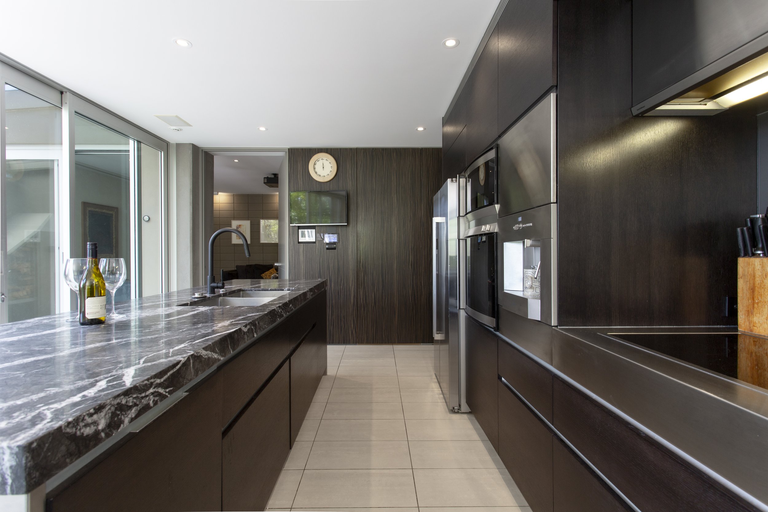 Modern kitchen with dark wood cabinets, stainless steel appliances, marble countertop island with wine and glasses, and a wall clock above a TV.