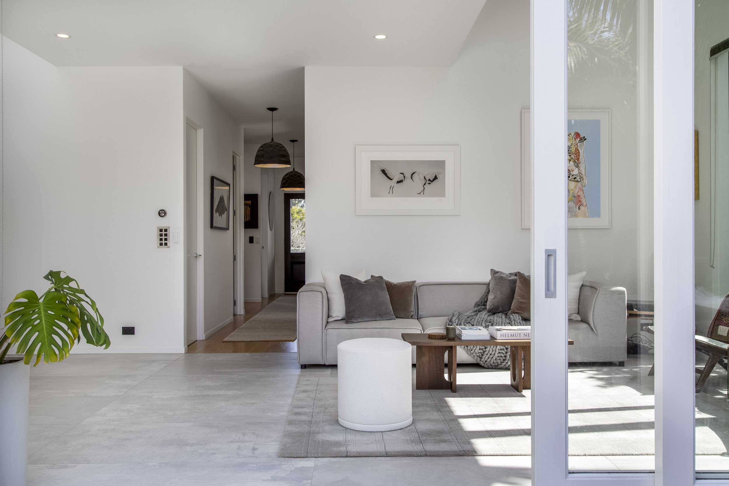 Modern living room with white walls, a light-colored sofa with gray pillows, a small round ottoman, and a wooden coffee table holding books and candles, with framed artwork on the walls, a sliding glass door revealing outdoor greenery, and a potted plant in the foreground.