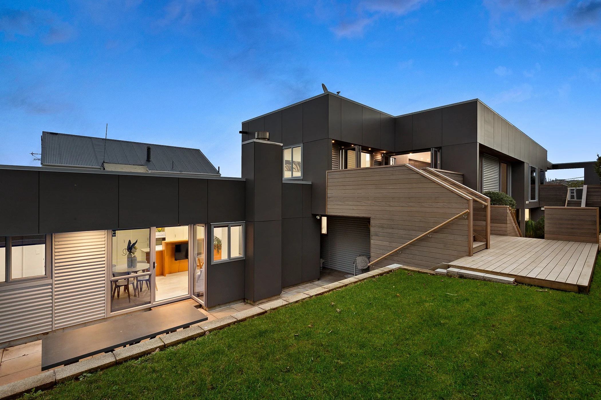 Modern multi-story house with black and wooden exterior, large windows, and an outdoor wooden deck with steps leading up to it, under a partly cloudy sky.