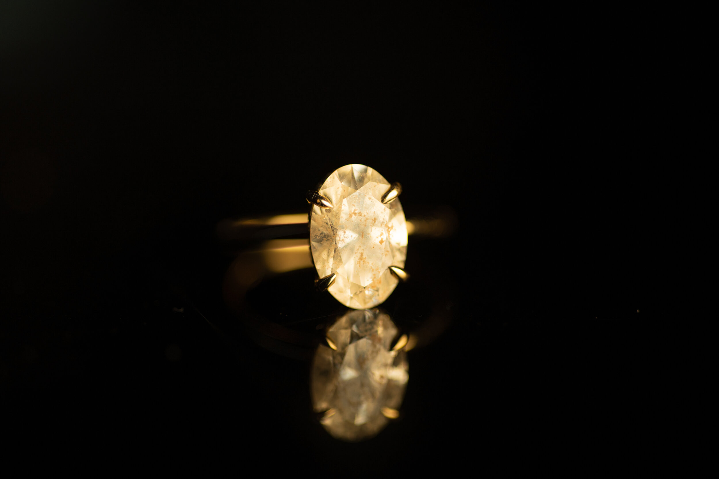 A gold ring with a large oval diamond gemstone against a black background, with its reflection visible beneath.