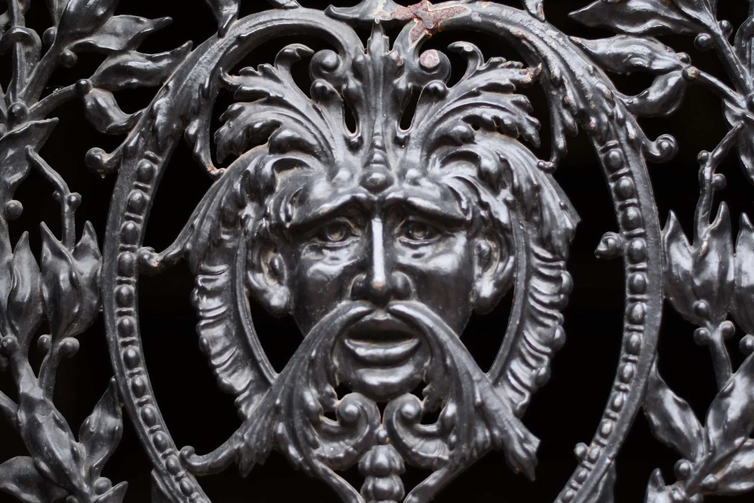 Close-up of an ornate black metal gate with a face in the center surrounded by decorative leaf patterns. Memphis architectural photography.