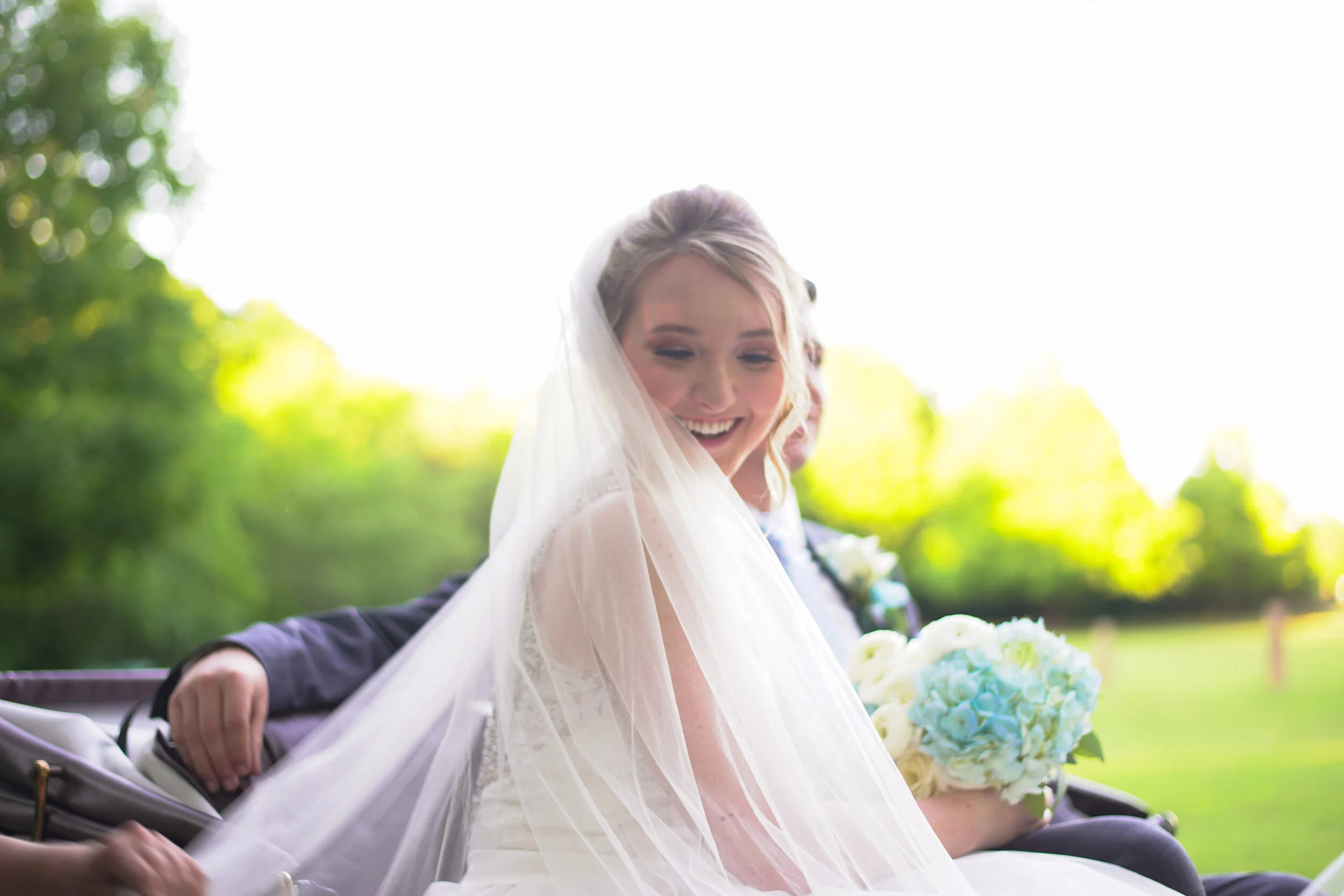 A bride smiling in a white wedding dress and veil, holding a bouquet of white and blue flowers, sitting in a carriage or similar vehicle outdoors with a green, blurry background and sunlight.