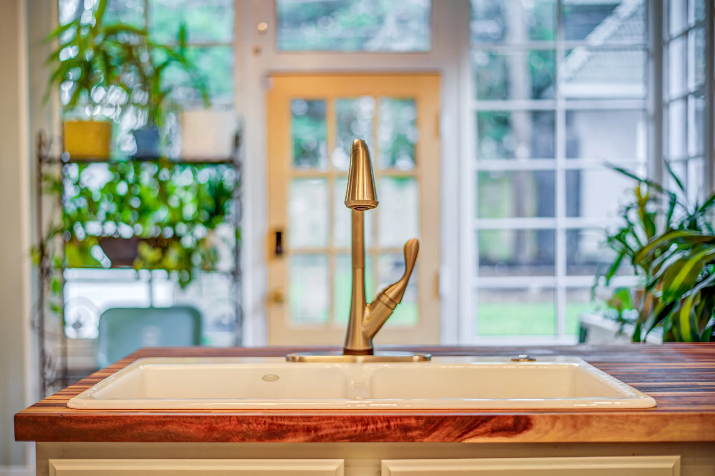 Kitchen sink with brushed gold faucet on a wooden countertop, with large windows and greenery outside in the background.