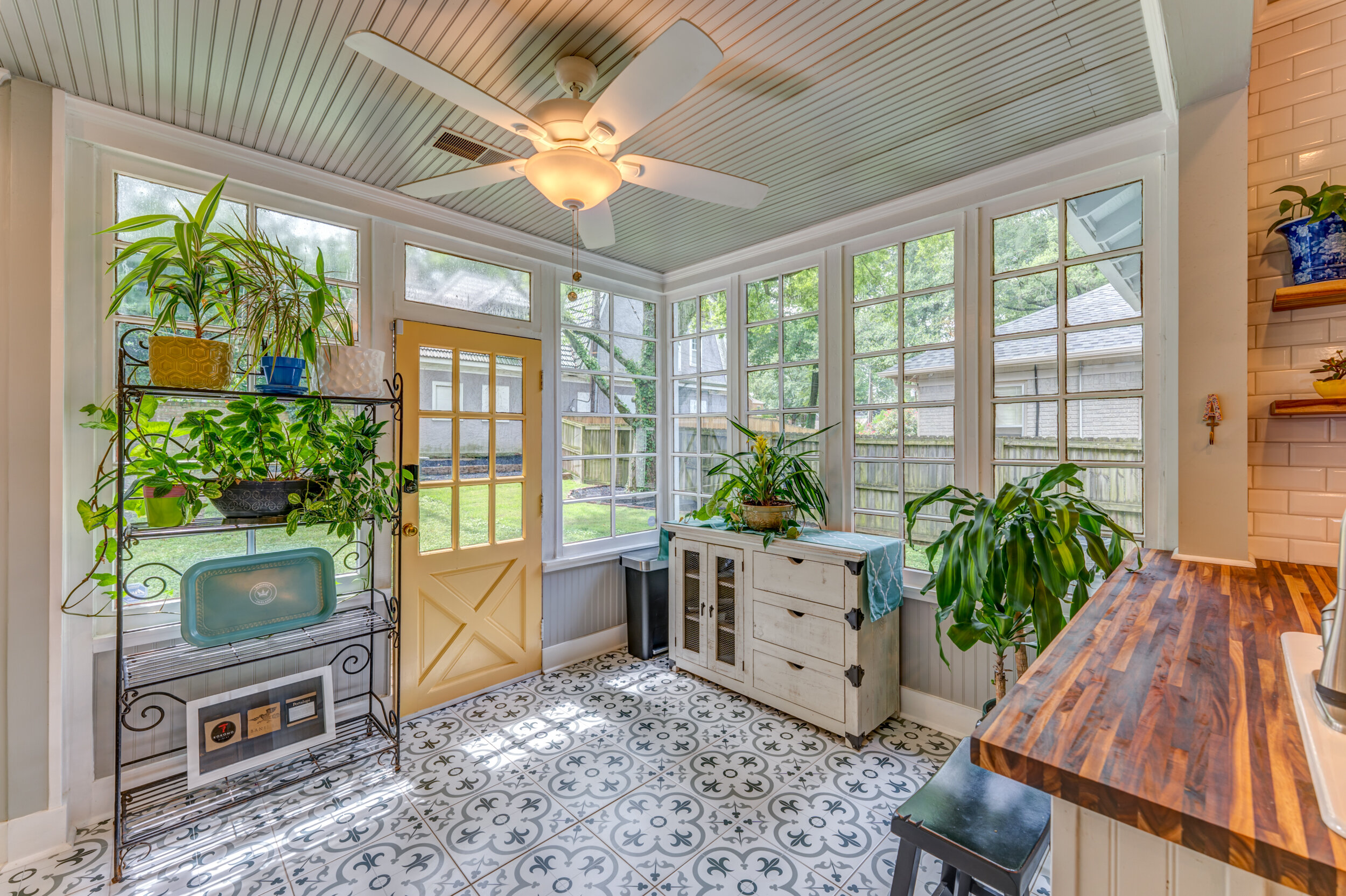 Bright enclosed porch with large windows, potted plants, a ceiling fan, and a patterned tile floor. Memphis real estate photography. 