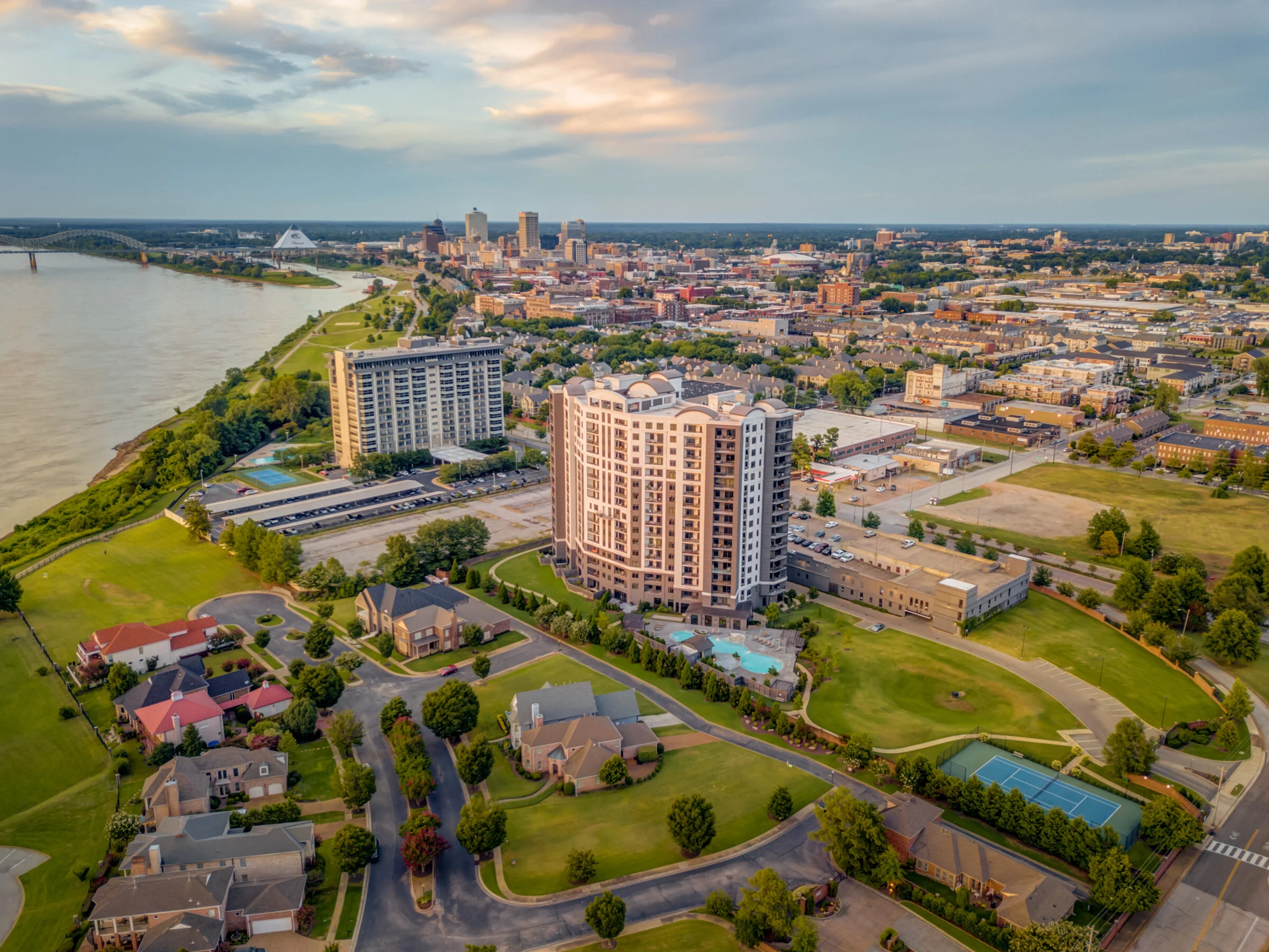 Aerial view of a city with a river on the left, high-rise buildings in the foreground, residential houses, golf course, tennis courts, and a downtown skyline in the background at sunset.